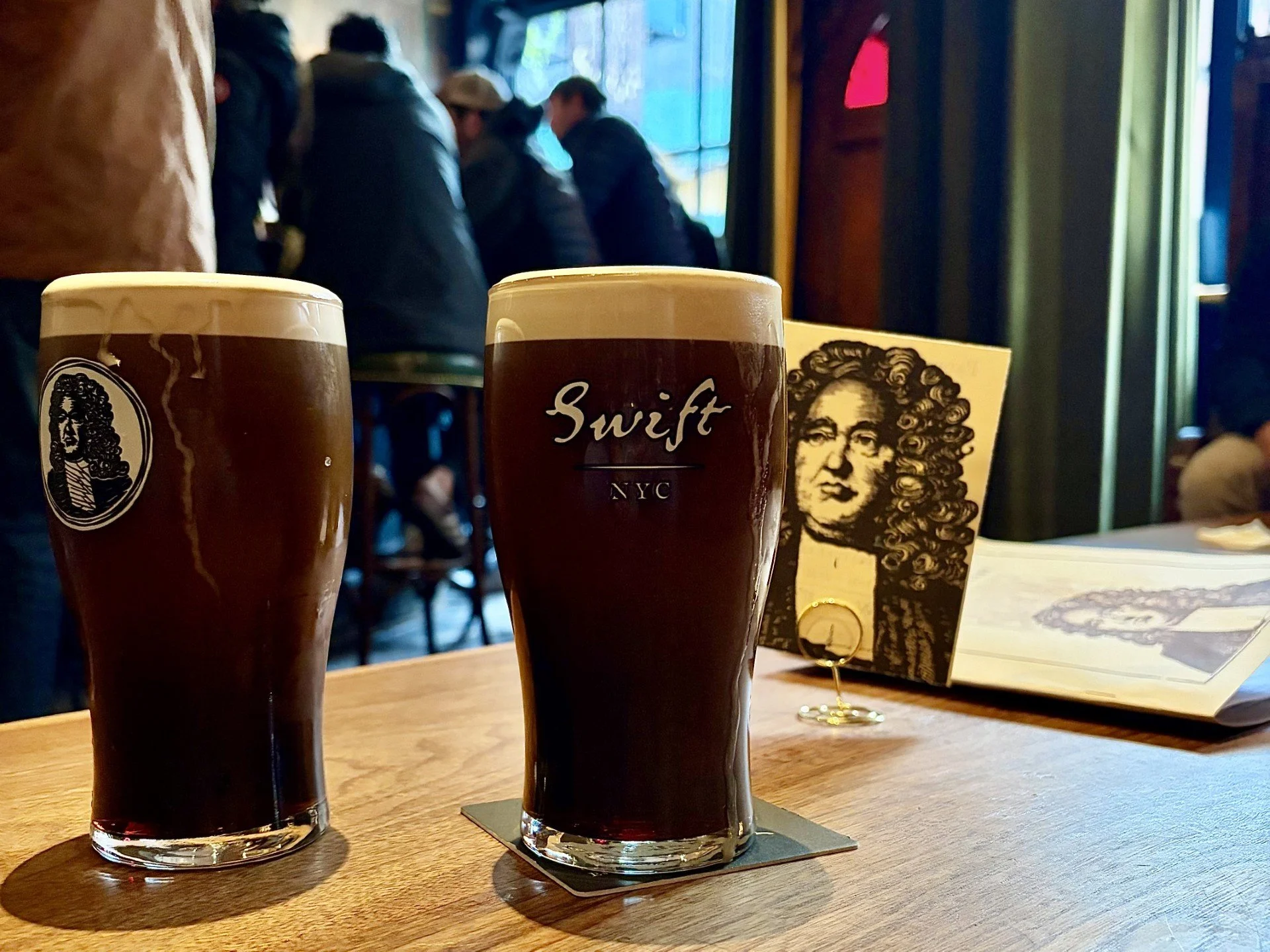 Two pints of Guinness on a table with an image of Jonathan Swift behind them.
