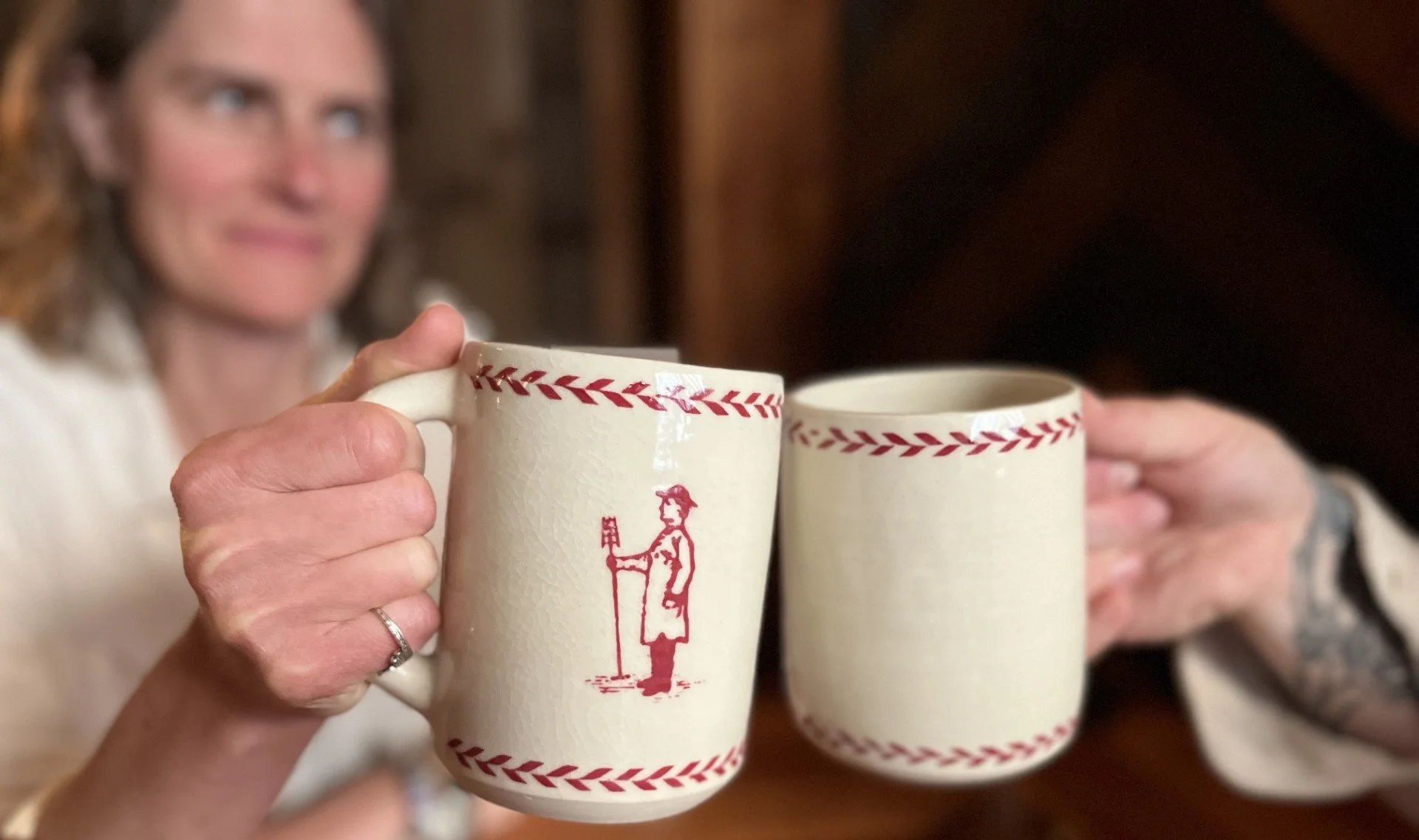 Two ceramic mugs being toasted with an out of focus woman in the background. The mug has an image of a brewer with a mash tun paddle.
