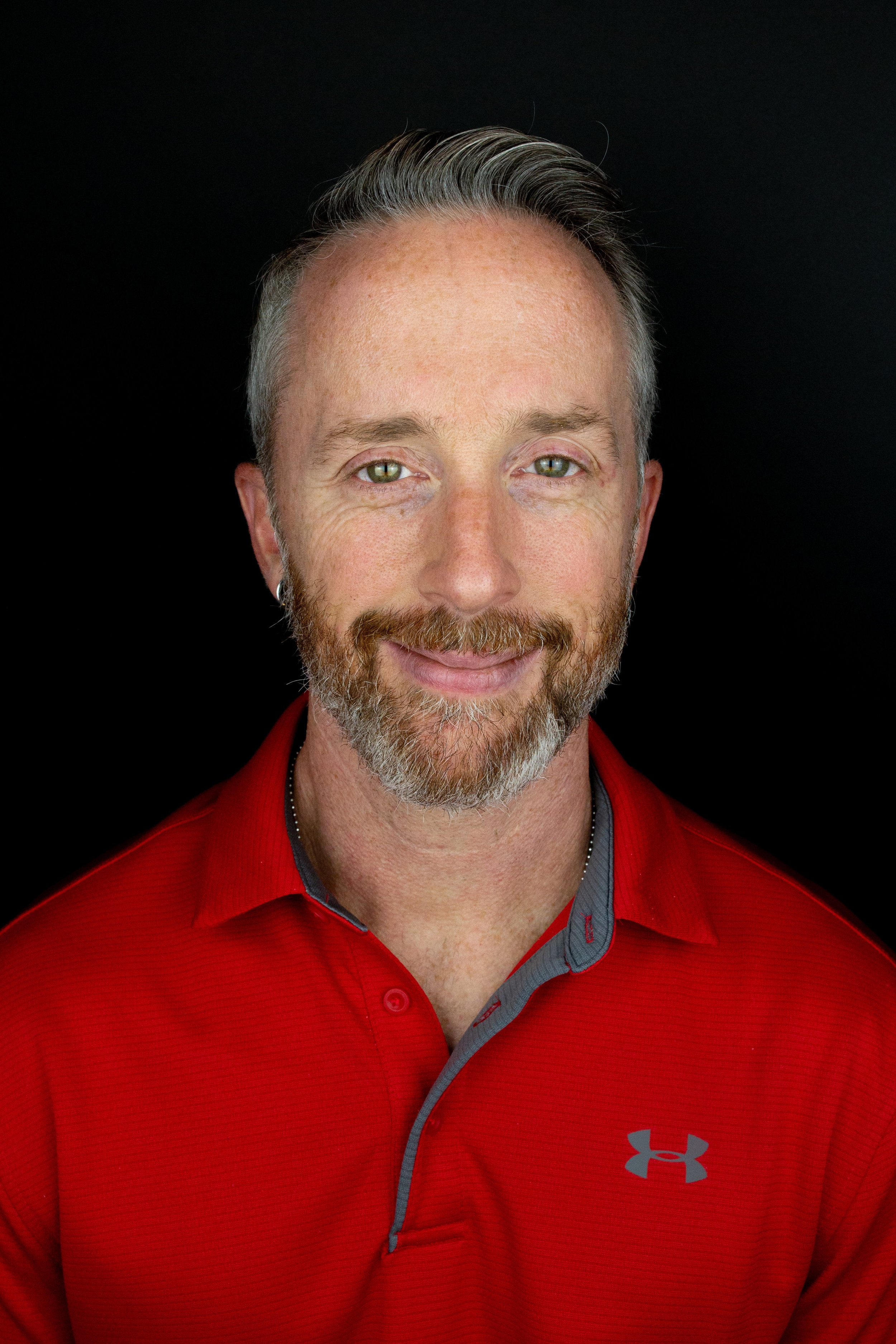 A man with light skin, gray hair, and a beard, smiling, wearing a red Under Armour polo shirt, against a black background.