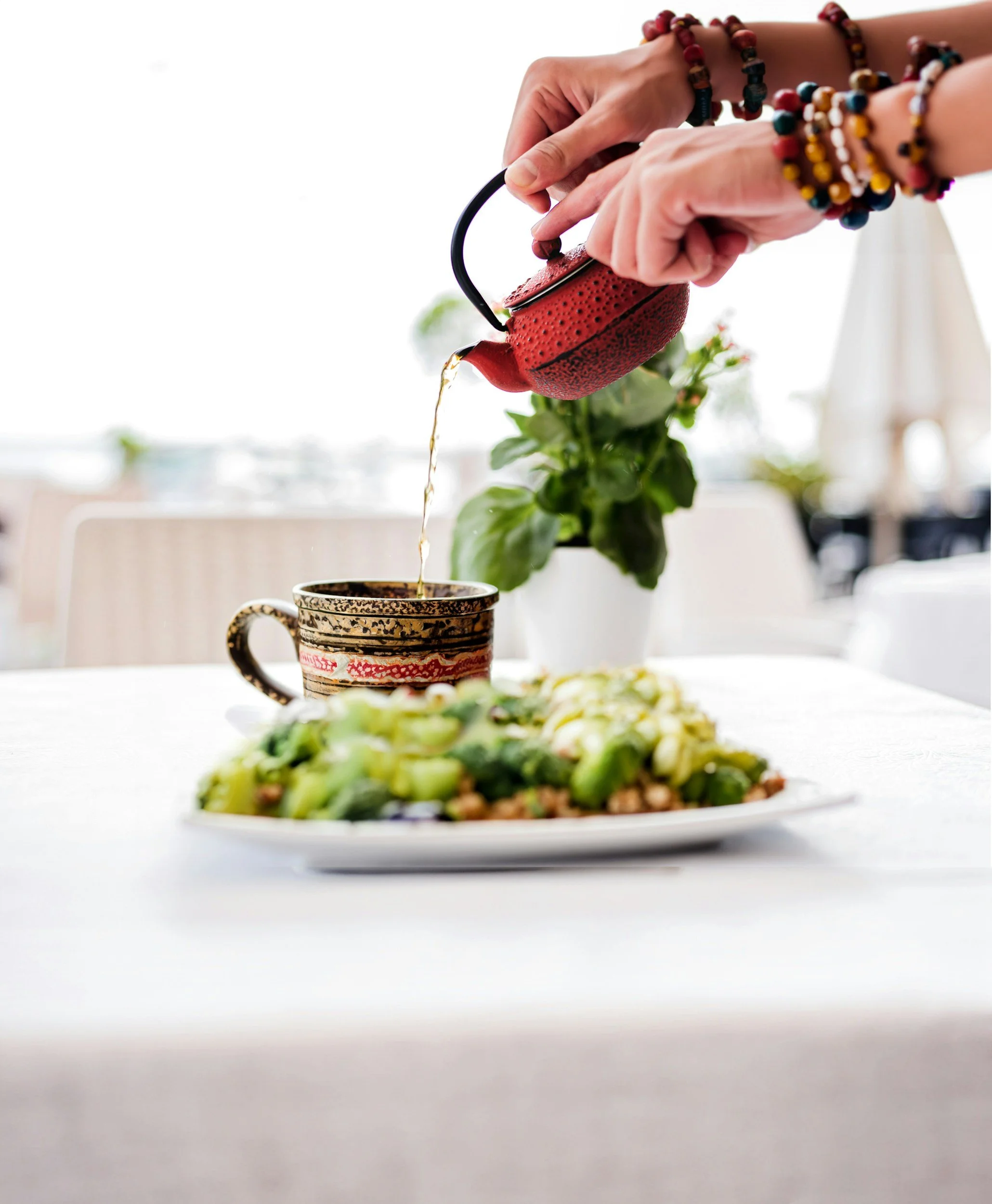 Person pouring tea from a red teapot into a decorated mug on a table with a leafy plant in the background and a plate of green vegetables in the foreground.