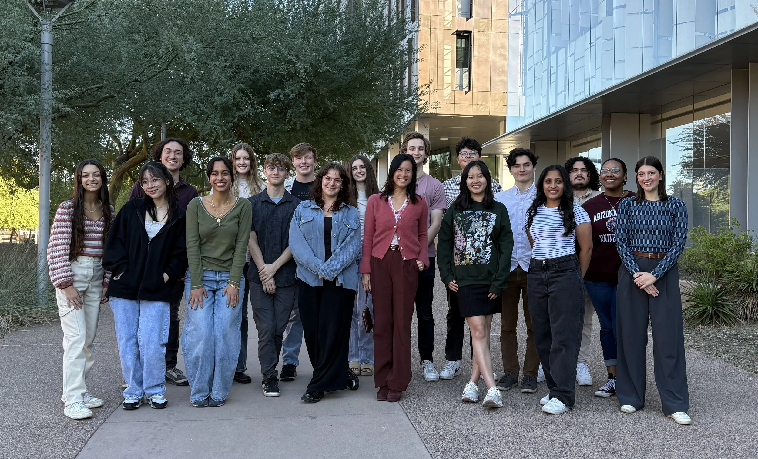 A diverse group of young adults standing outdoors, smiling, in front of a modern glass building with trees in the background.