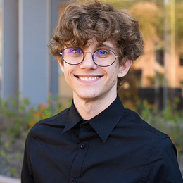 Young man with curly brown hair, wearing round glasses and a black button-up shirt, smiling outdoors with modern buildings and greenery in the background.
