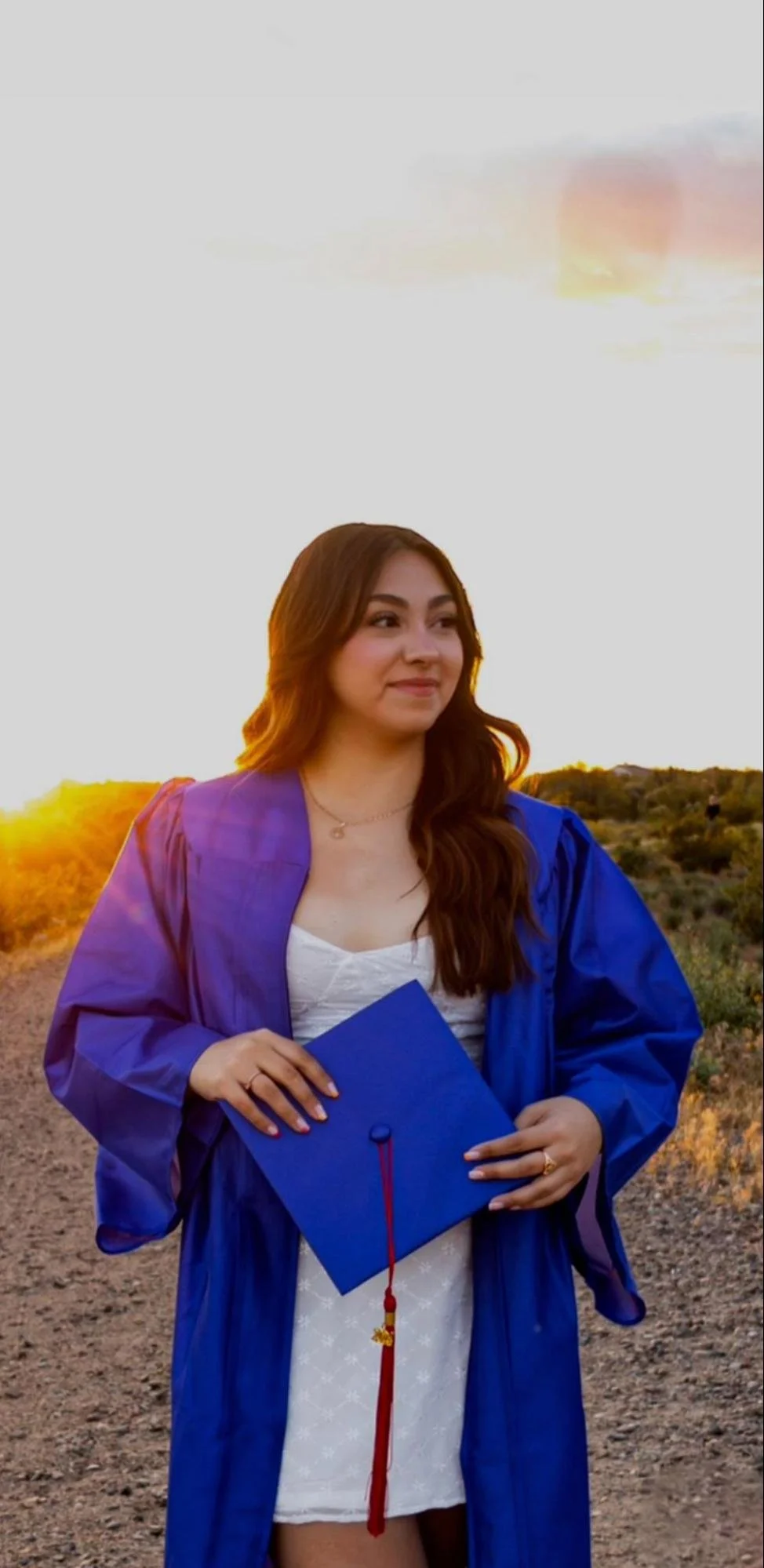 A woman in a blue graduation gown holding a blue mortarboard cap standing outdoors during sunset with a landscape of sparse bushes and a dirt path.