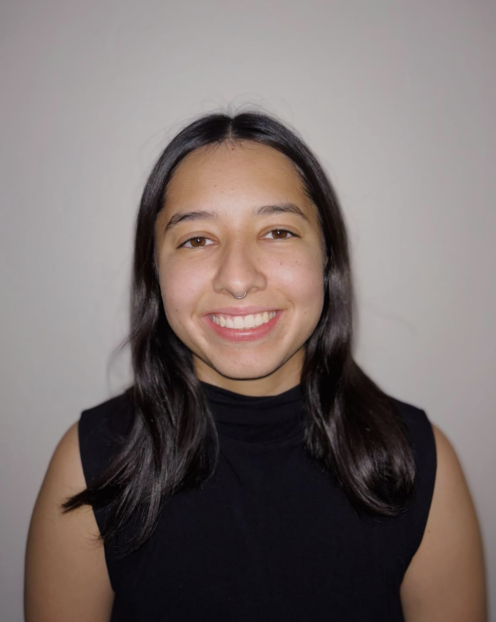 A young woman with shoulder-length brown hair smiling, wearing a septum piercing, a white t-shirt, and a gray vest, standing against a beige textured wall.