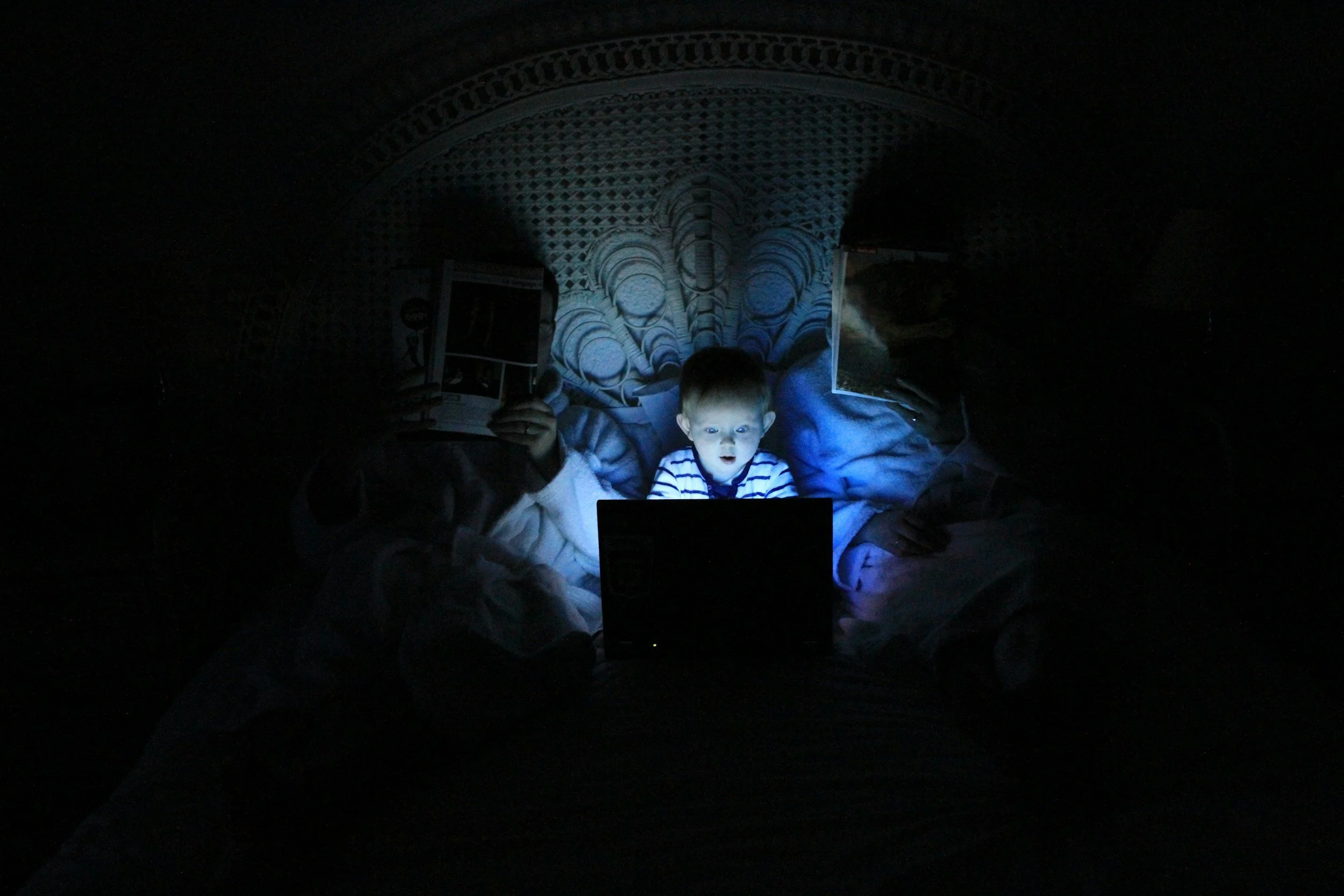 A young boy is lying in bed in a dark room, illuminated by the glow of a laptop screen, with a person beside him holding a book.