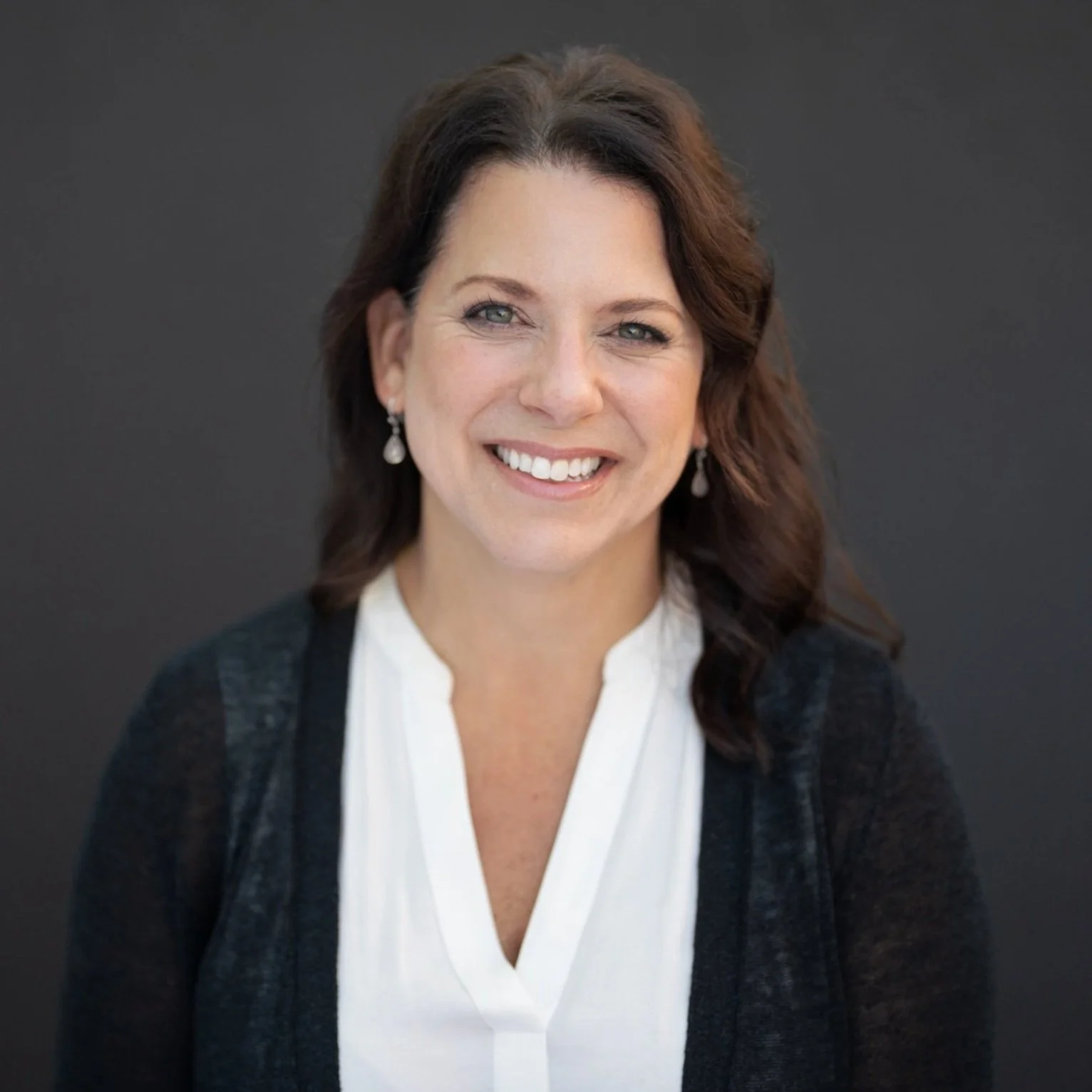 Portrait of a woman with brown hair, blue eyes, white blouse, black cardigan, smiling against a dark background.
