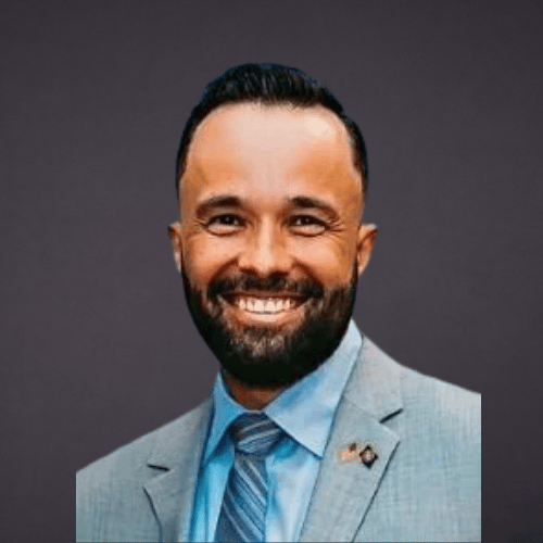 Portrait of a smiling man in a light gray suit, blue dress shirt, and striped tie, against a dark background.