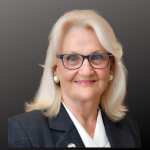 A woman with blonde hair wearing glasses, pearl earrings, a dark blazer, and smiling against a dark background.