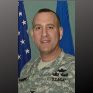 A man in U.S. Army military uniform standing in front of American and blue flags.