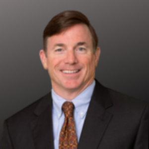 A professional man wearing a suit and tie, smiling against a gray background.