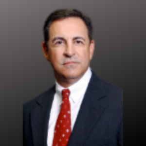 A man in a dark suit with a white shirt and red polka dot tie, posing against a plain dark gray background.