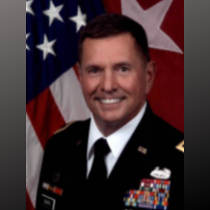 Portrait of a man in military uniform with medals, standing in front of an American flag and another flag.