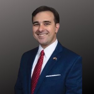 A man in a navy blue suit, white shirt, and red dotted tie smiling against a gray background.
