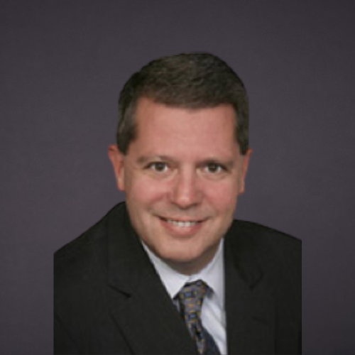 A professional headshot of a man wearing a black suit, white shirt, and patterned tie, smiling against a dark background.
