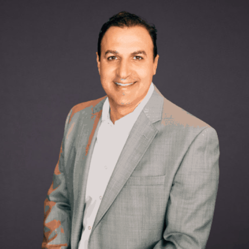 Professional portrait of a smiling man with dark hair in a gray blazer and white shirt against a dark background.