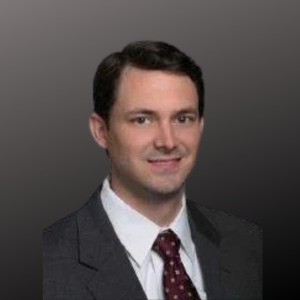 Professional headshot of a man in a suit and tie, smiling against a gray background.