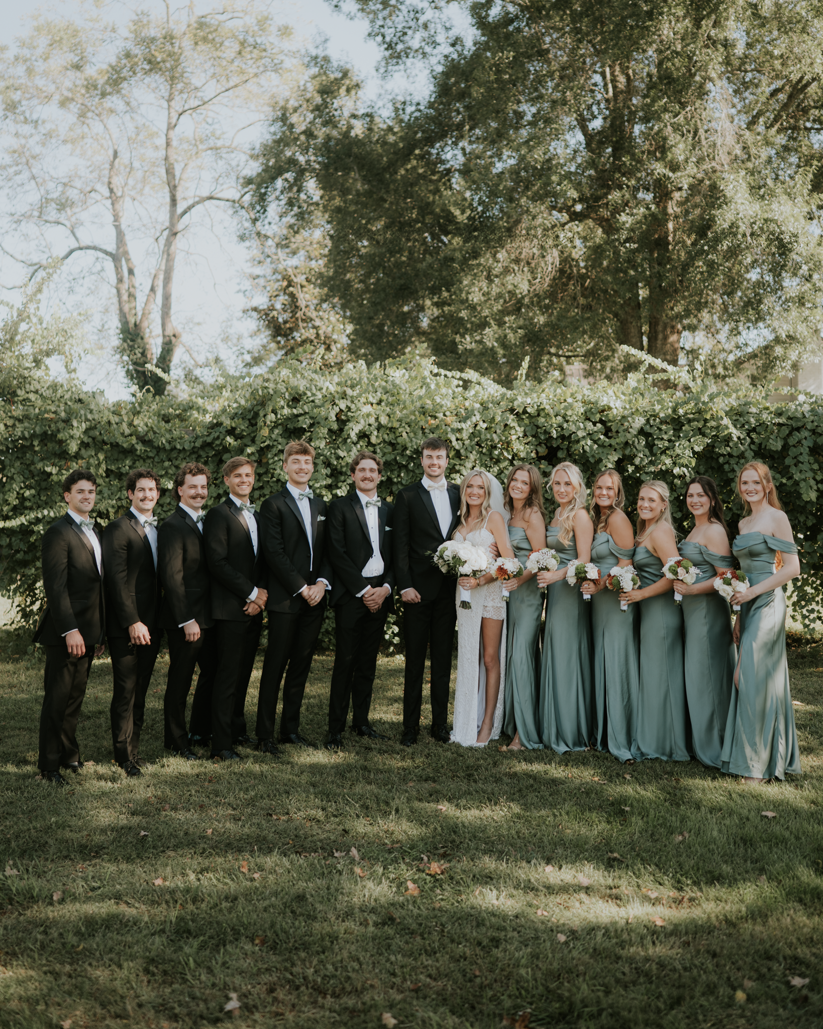 A group of people dressed in formal attire, with men in tuxedos and women in bridesmaid dresses, posing together outdoors in front of a large green hedge and trees on a clear day, likely for a wedding.