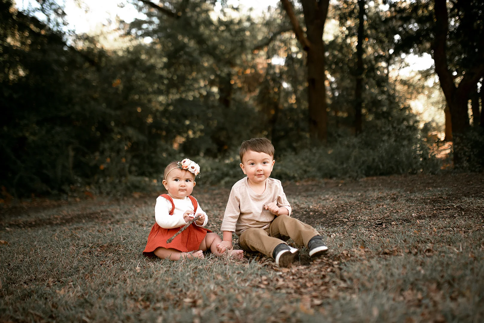Two young children, a girl and a boy, sitting on the ground outdoors in a wooded area at sunset. The girl is wearing a rust-colored dress with a white long-sleeve shirt underneath and a floral headband, holding a stick. The boy is in a beige shirt an