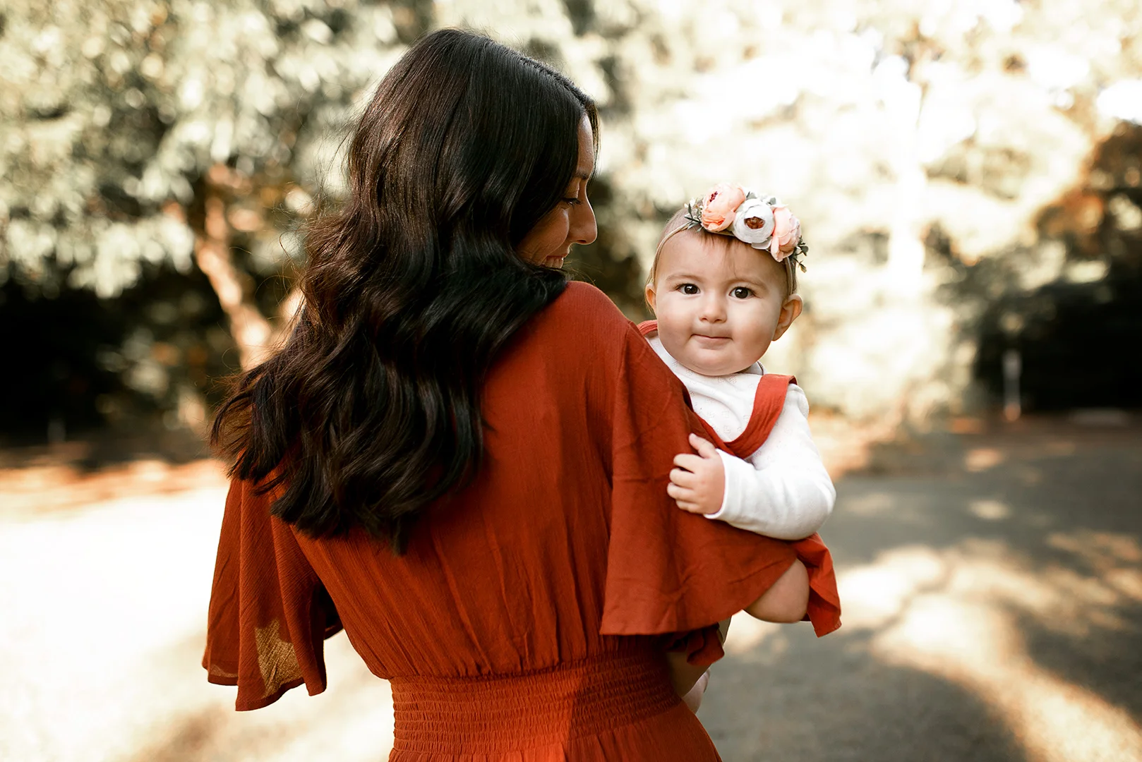 A woman with long black hair holding a baby girl outdoors surrounded by trees with autumn leaves. The woman is wearing a rust-colored dress; the baby has a floral headband and a white and rust-colored outfit.