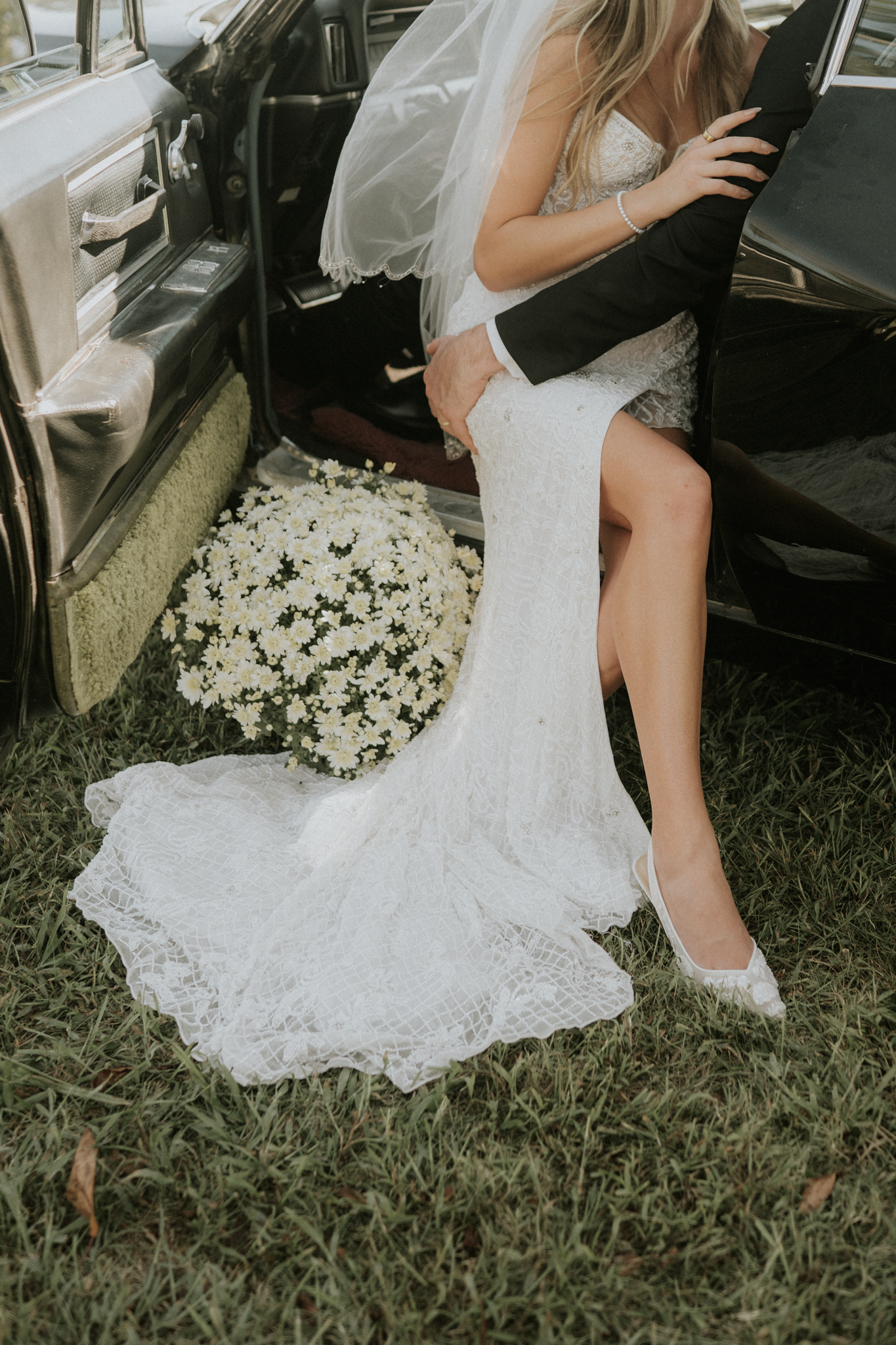A bride and groom kissing near an open vintage car with a large bouquet of white daisies on the ground beside them.