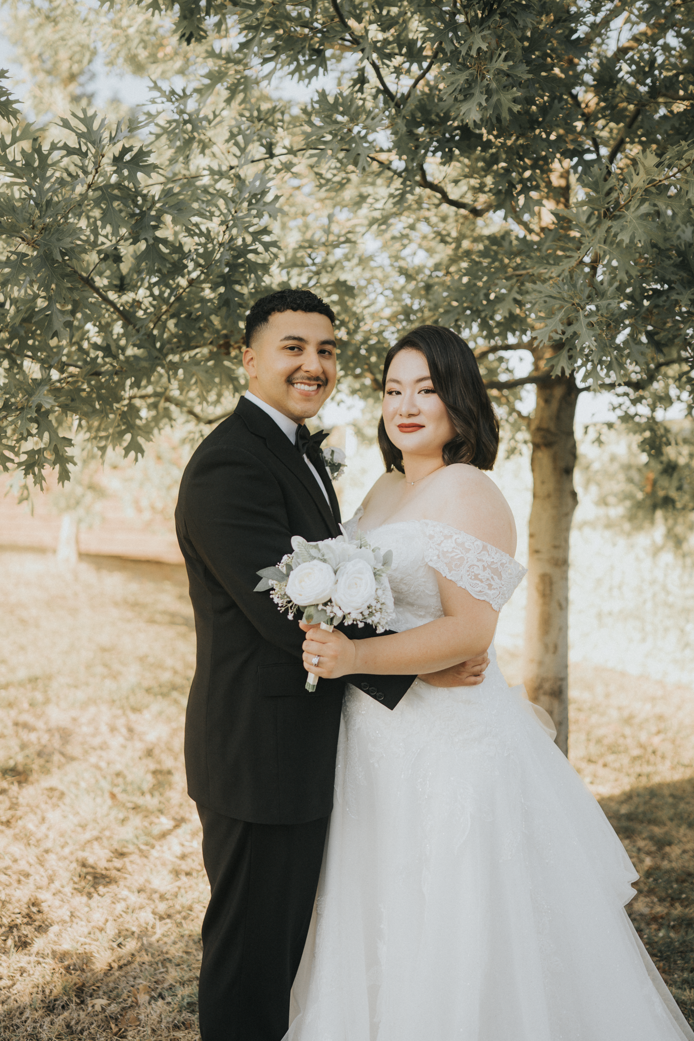 A newlywed couple stands under a leafy tree outdoors, with the groom in a black tuxedo and the bride in a white lace off-the-shoulder wedding gown. The groom is smiling, and the bride is holding a bouquet of white roses, with a serene expression.