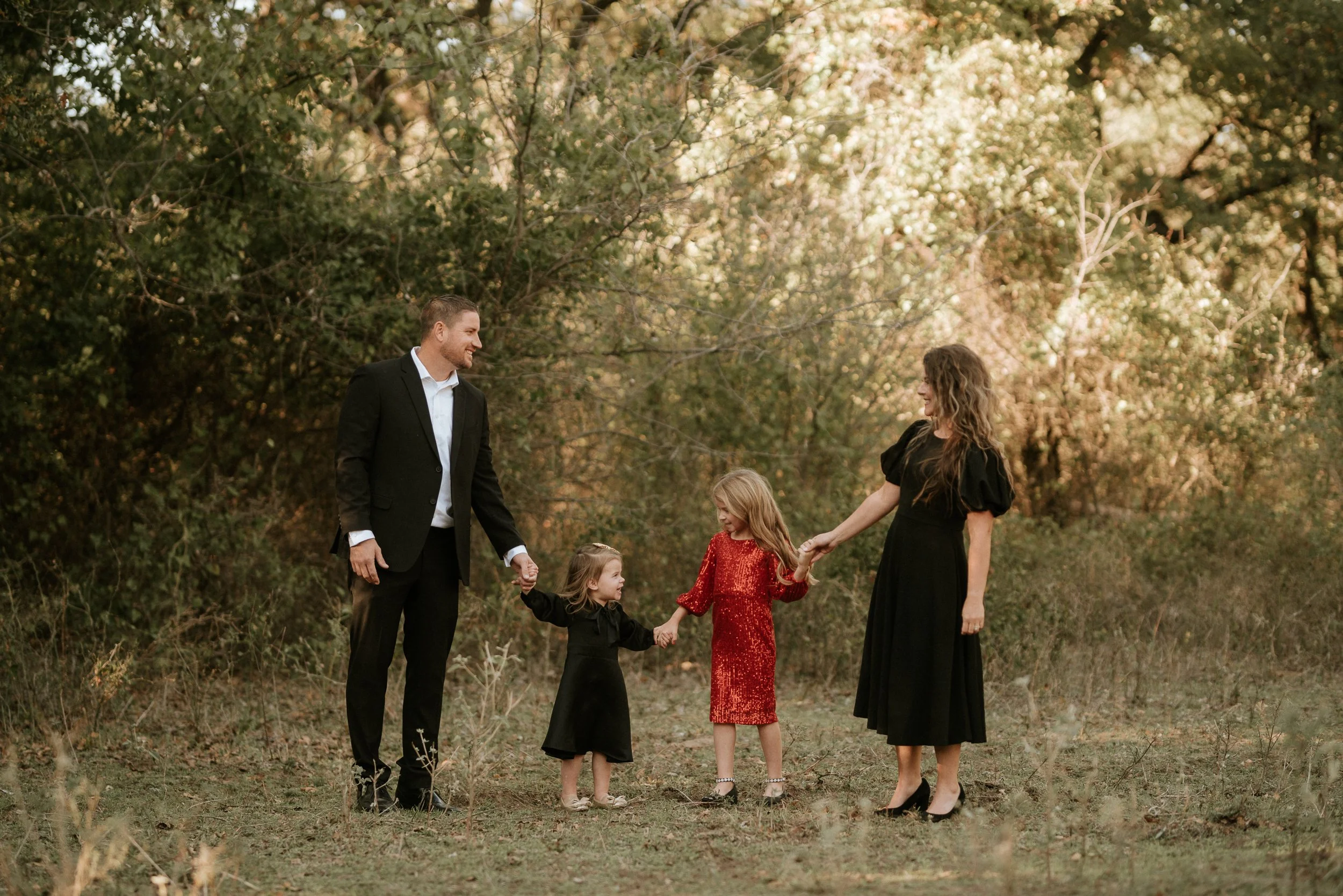 A family of five holding hands and standing in a wooded outdoor area during daytime. The father is wearing a suit, the mother is in a black dress, and three young girls are dressed in red and black. They are smiling and looking at each other, enjoyin