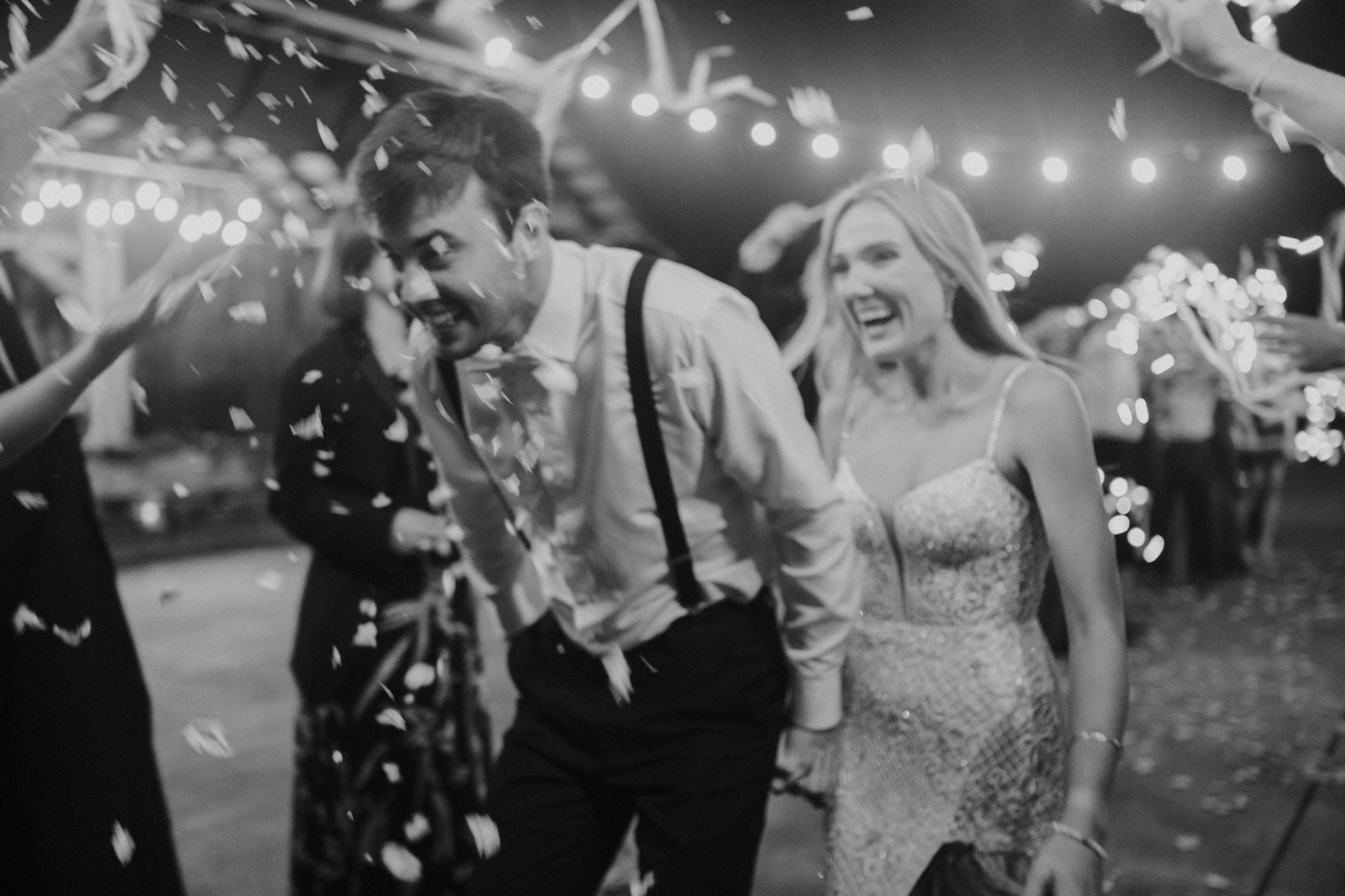 A black and white photo of a couple at a celebration, possibly a wedding, surrounded by falling confetti and string lights. The man, wearing suspenders and a white shirt, is smiling and looking to his right. The woman, in a lace dress, is smiling and