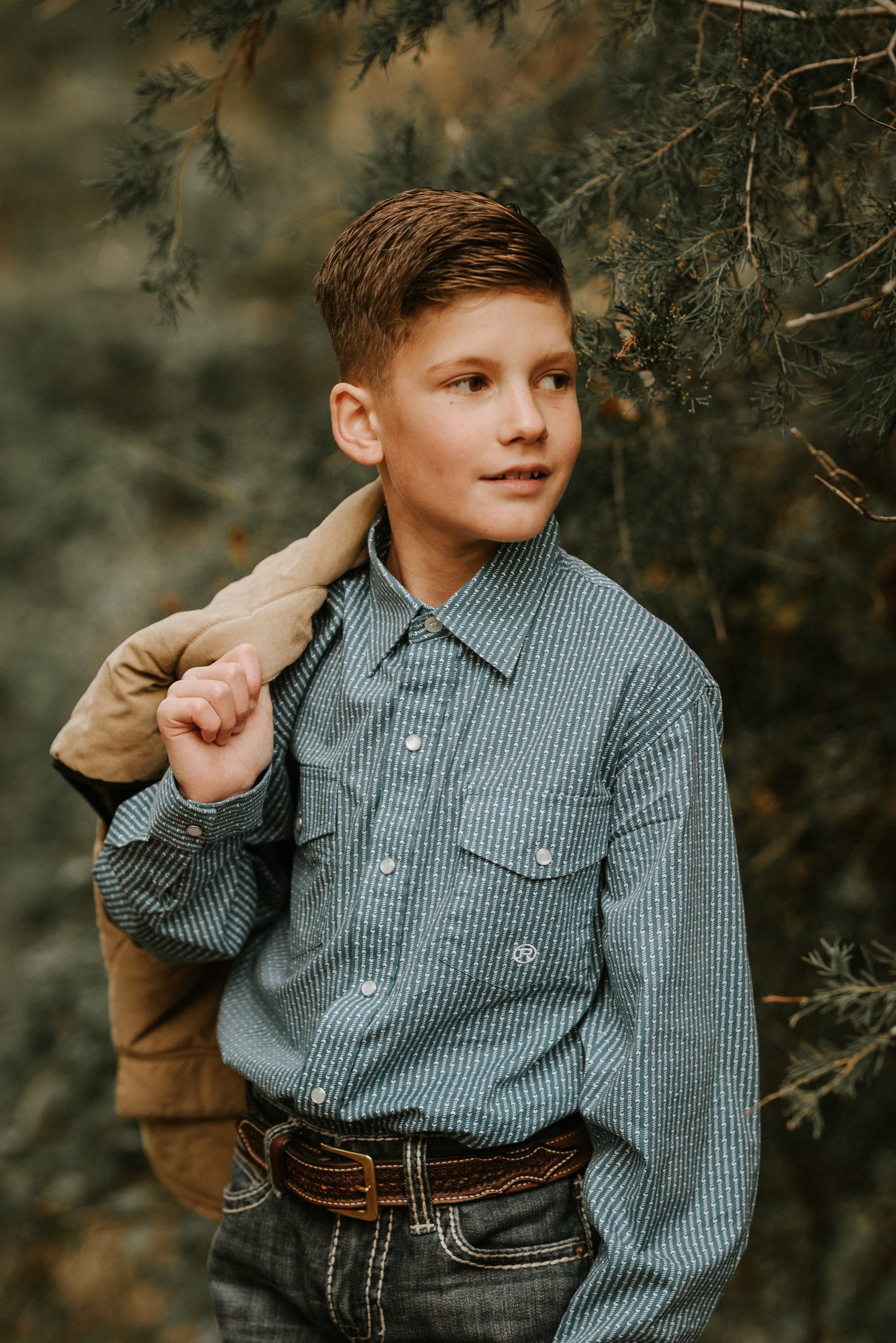 A young boy with short brown hair wearing a button-up shirt with thin vertical stripes, dark jeans, and a brown belt is standing outdoors among evergreen branches. He has a tan jacket draped over his shoulder and is looking to his right.