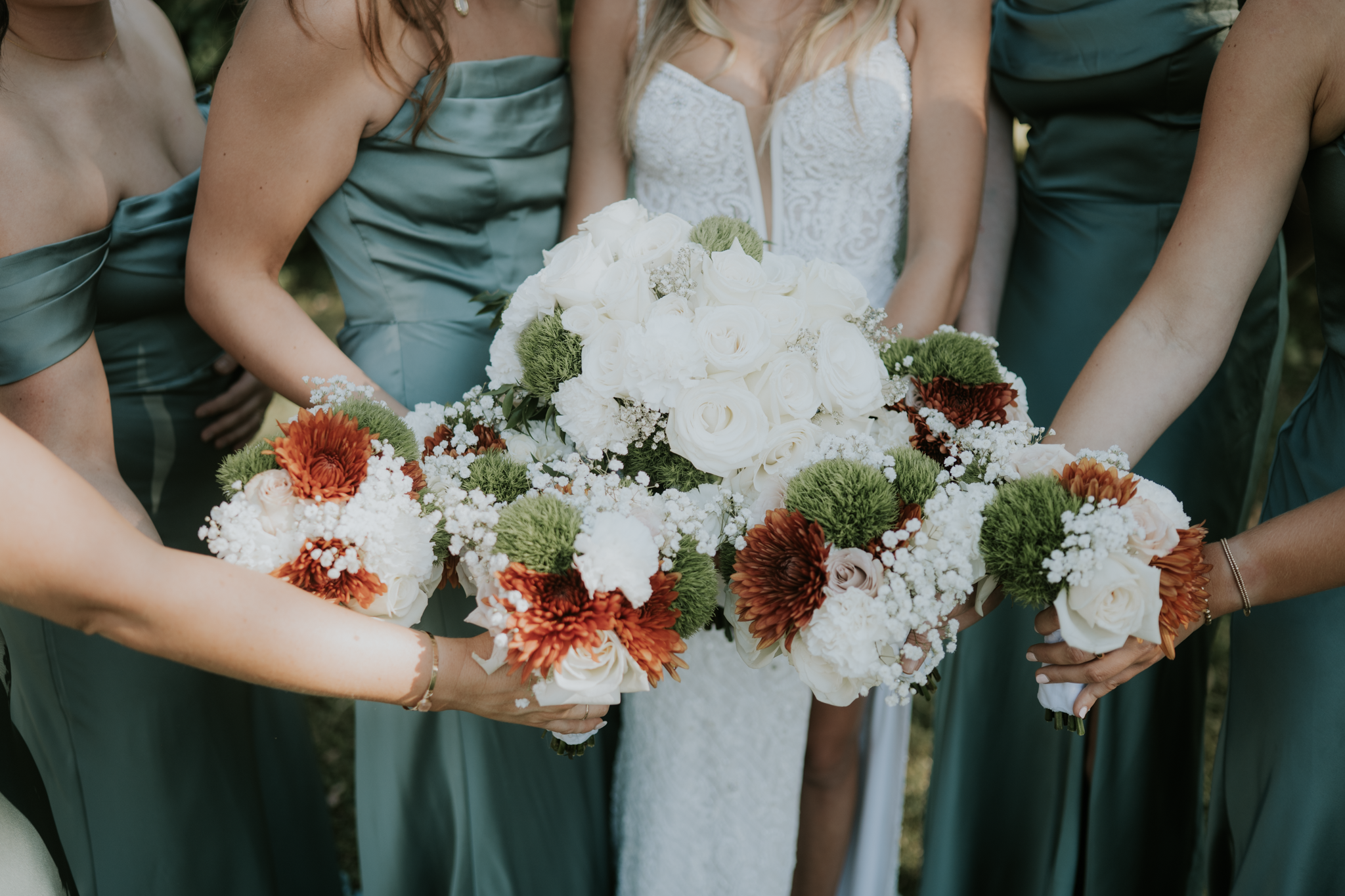 A bride in a white wedding dress holding a bouquet of white roses and greenery, surrounded by bridesmaids in teal dresses holding their own bouquets of flowers.
