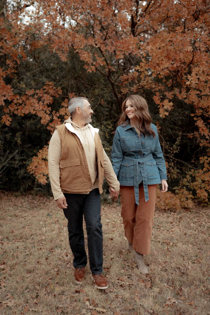 A middle-aged man and a young woman hold hands while walking in a park during autumn, surrounded by orange and red maple trees.