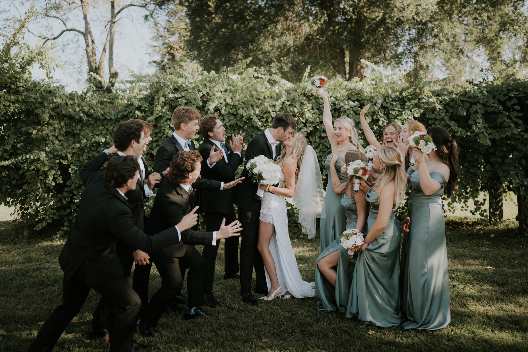 A wedding celebration outdoors with the bride and groom kissing, surrounded by bridesmaids and groomsmen cheering and holding bouquets, against a backdrop of green foliage.