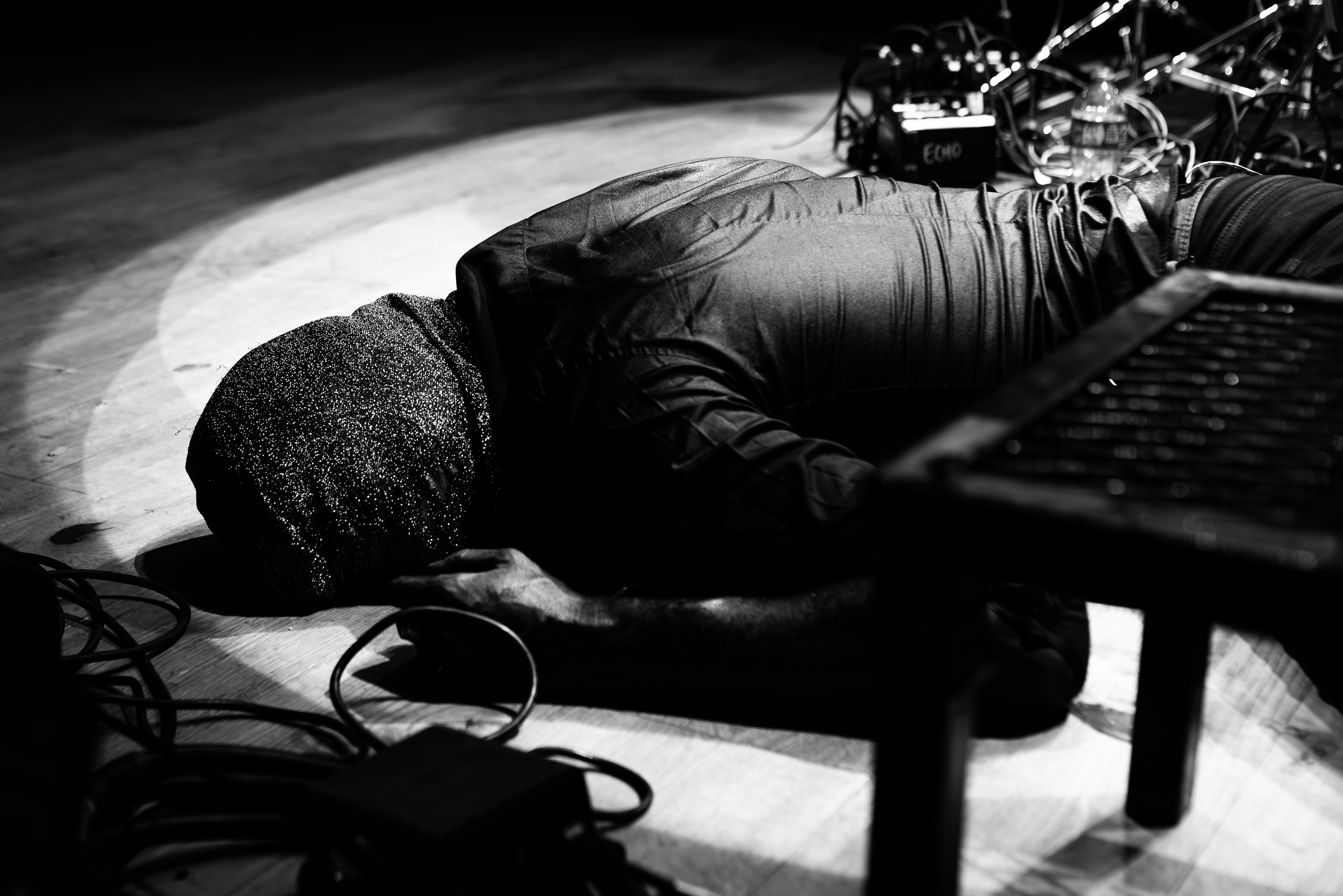 A person lying face down on the floor with wires and electronic equipment around them, a water bottle near their head, in a black-and-white photo.