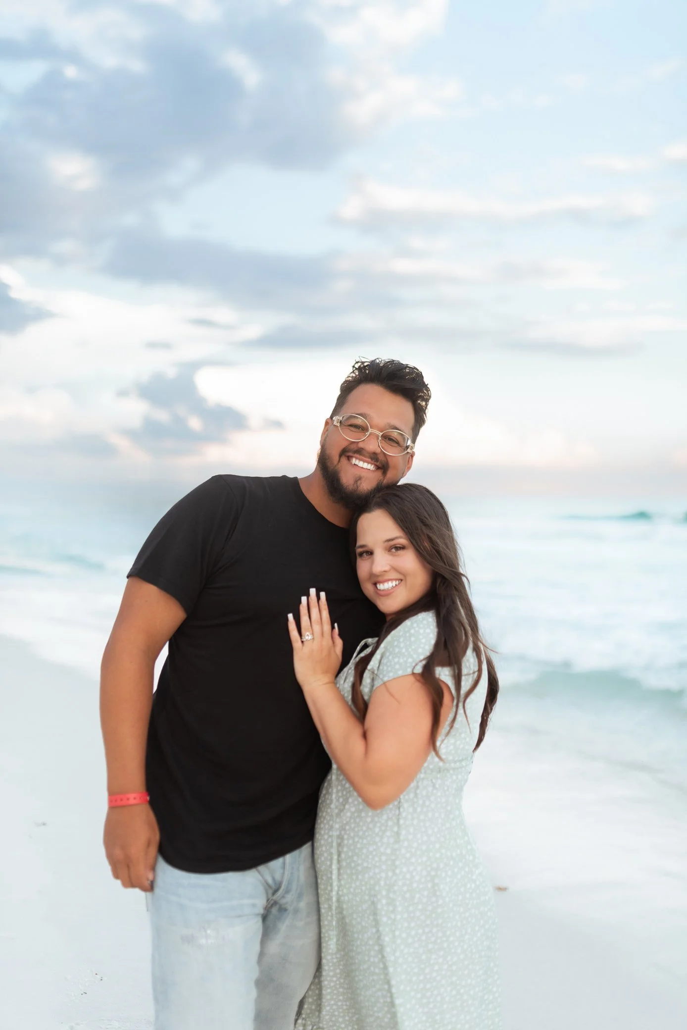 A smiling man and woman standing on a beach, the man wearing glasses and a black T-shirt, the woman with long brown hair in a light green dress, both showing the woman's engagement ring, with the ocean and sky in the background.