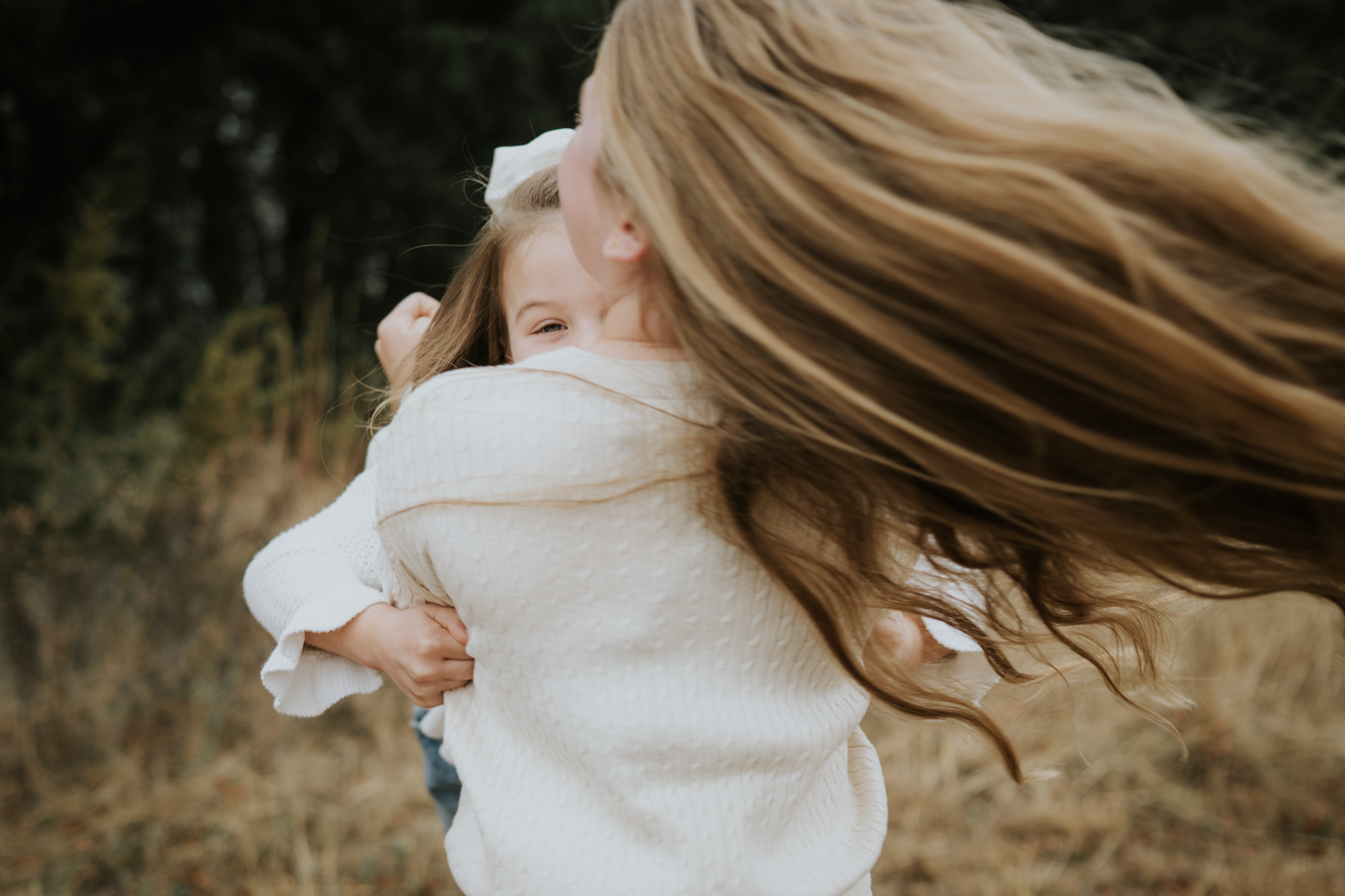 A woman with long red hair hugging a smiling girl with light brown hair outdoors in a field with trees in the background.