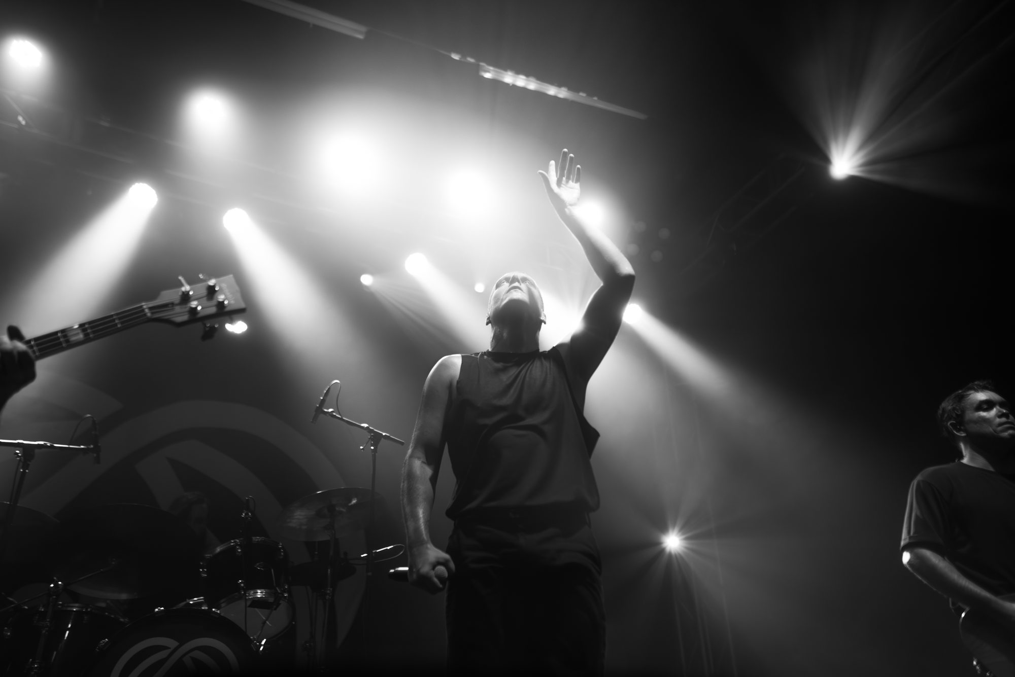 Black and white photo of a live music performance. A woman with braided hair and wearing a sleeveless top is raising her right arm, with stage lights shining behind her. A guitarist's headstock and part of a drum set are visible in the foreground, an