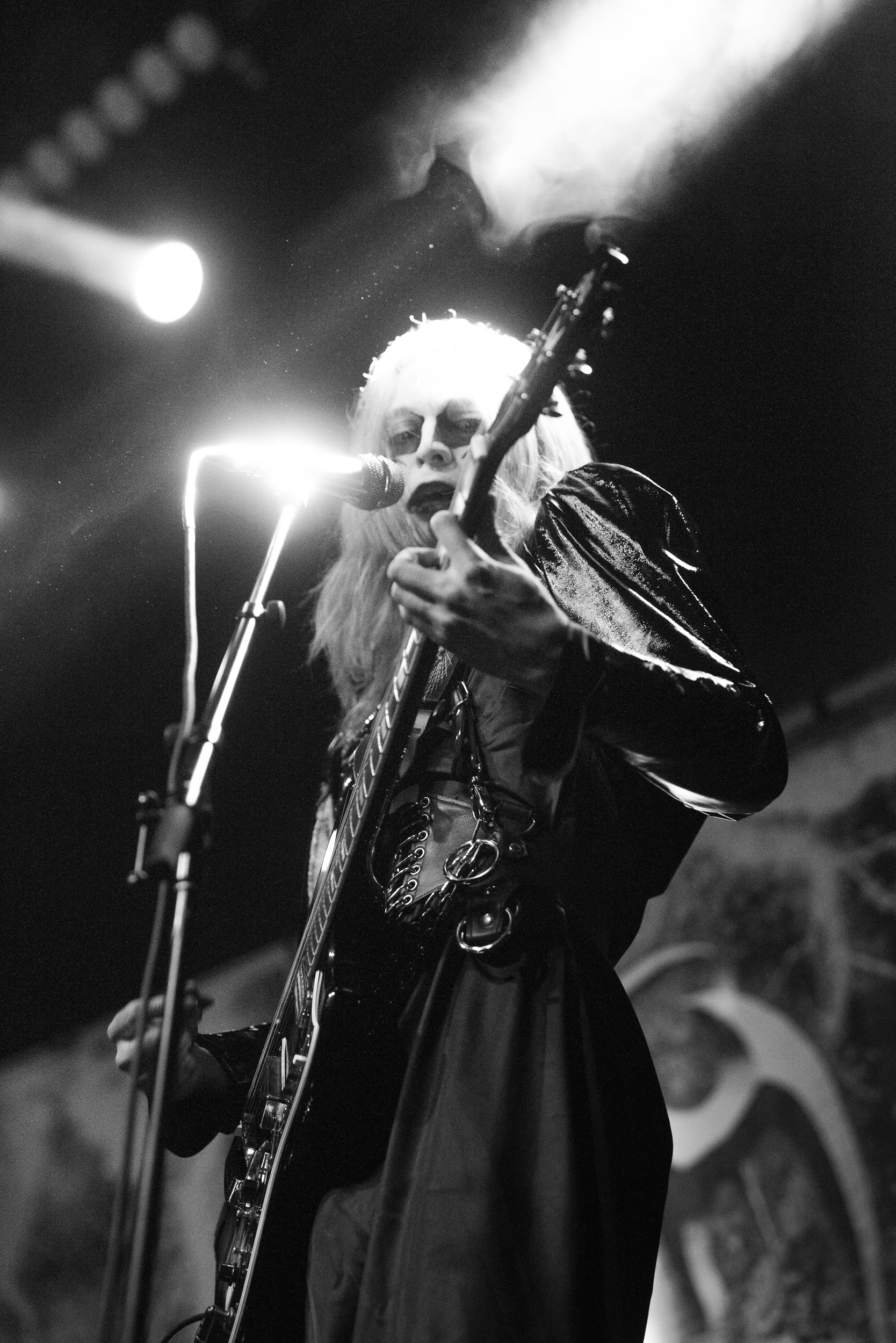 A black and white photo of a woman singing into a microphone while playing an electric guitar on stage. She has long hair and is wearing sunglasses and a shiny jacket.