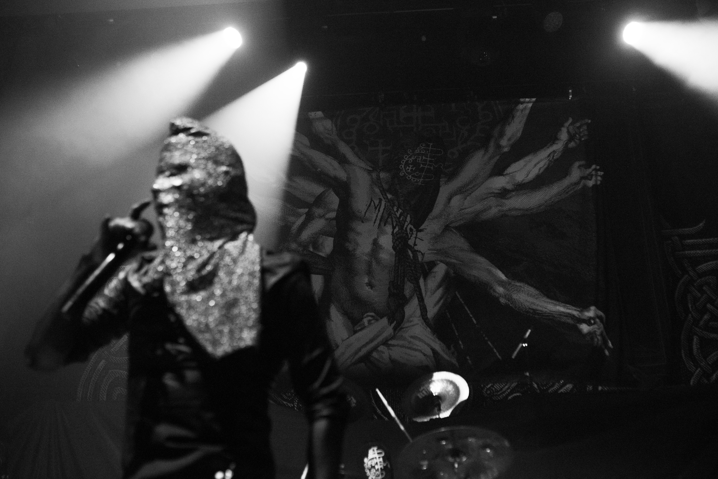 Black and white photo showing a person pointing a finger towards the camera with a distressed face. They are wearing a shiny, glittery garment. Bright lights are shining from above, illuminating the scene with a dramatic effect. In the background, th