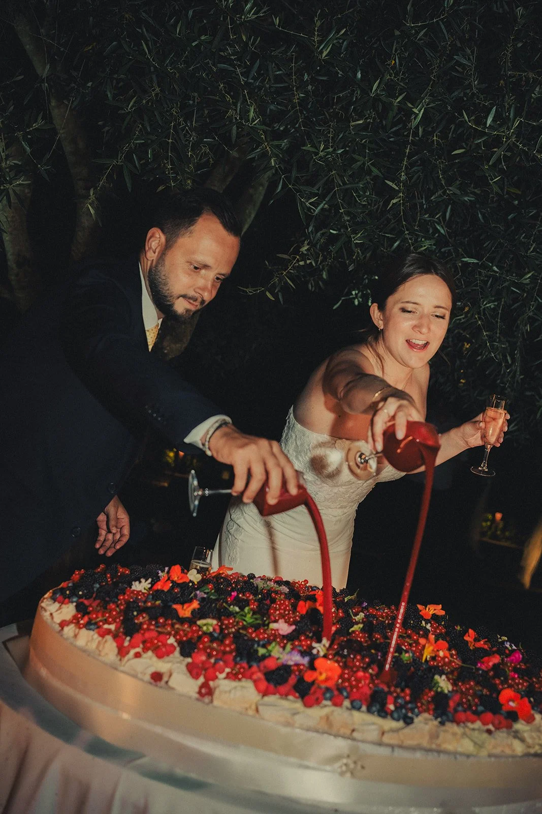 South of France wedding photographer capturing bride and groom pouring berry sauce on their large wedding cake full of berries.