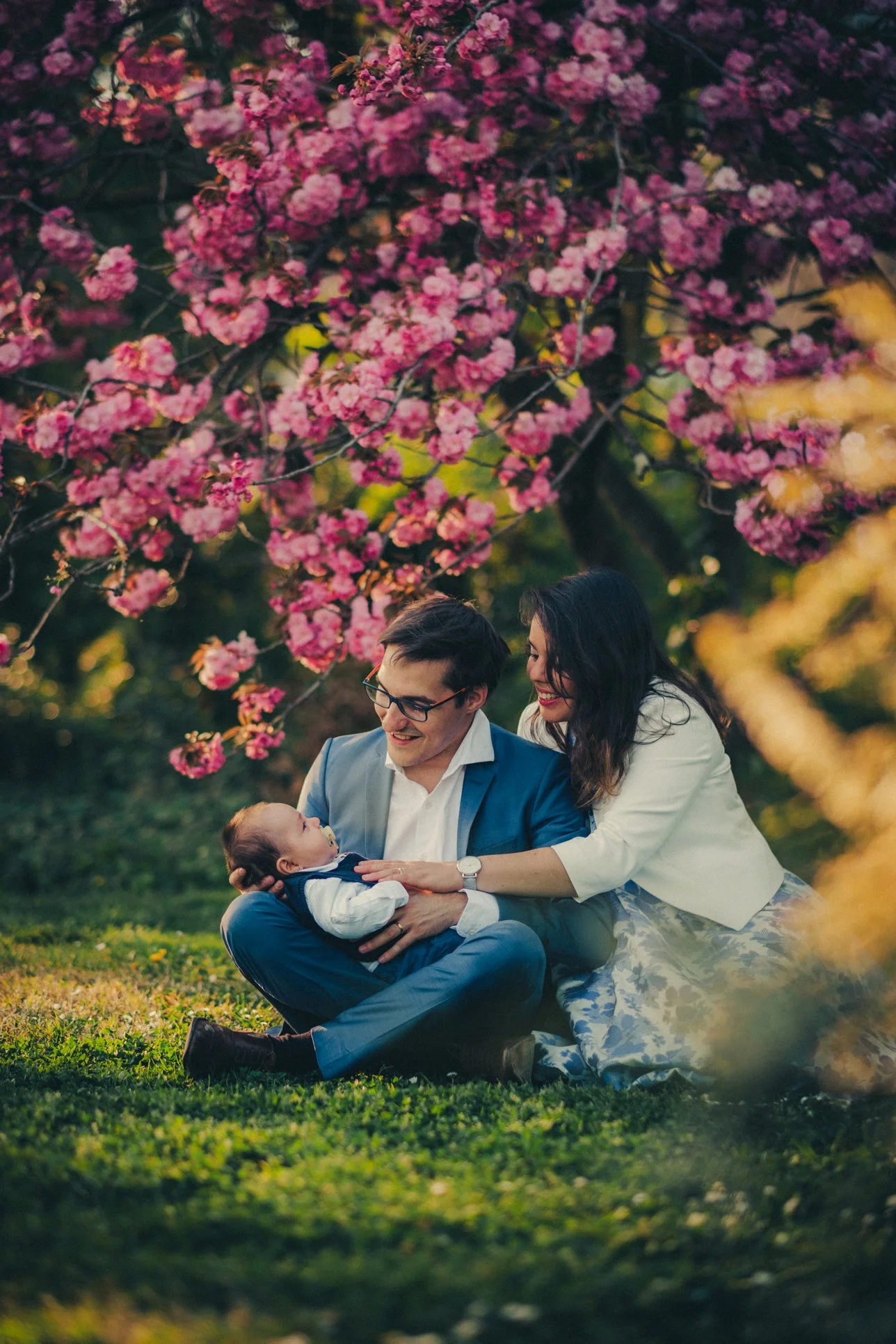 Parents avec leur nouveau-né assis sous les arbres en fleurs pendant une séance photo famille au parc de la Tête d’Or à Lyon