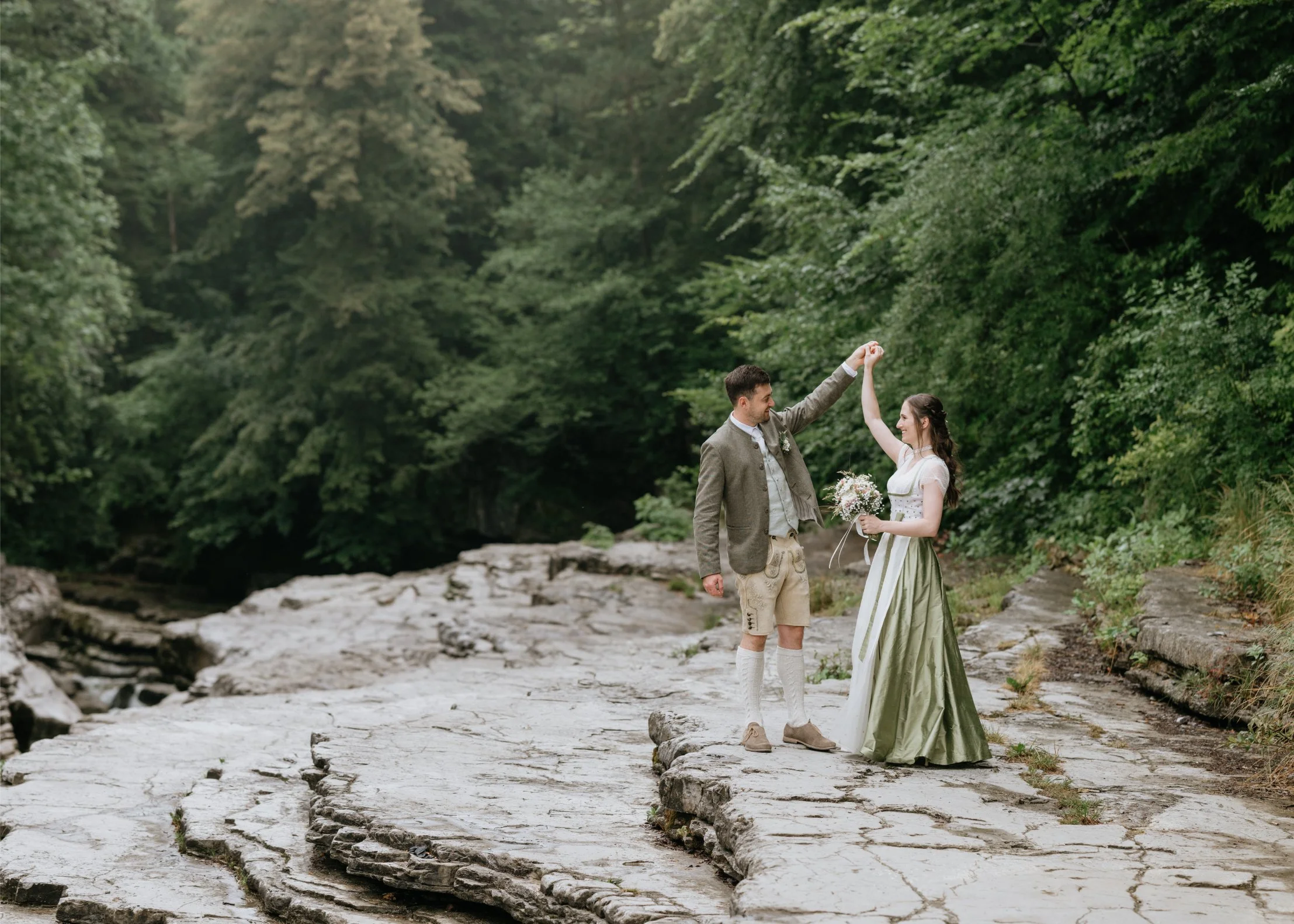 A man and woman dressed in traditional German or alpine attire, holding hands and smiling in a forested outdoor setting.