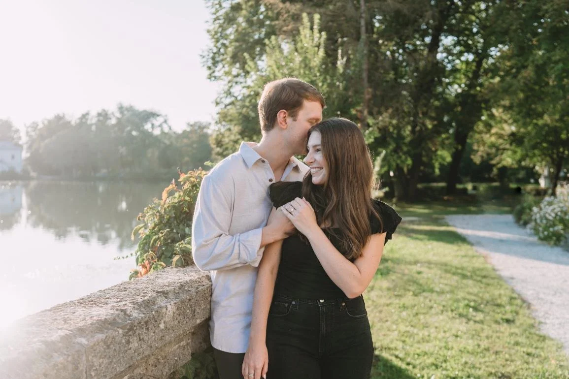 A young couple sharing a romantic moment by a lakeside in a park, with trees and a gravel path in the background.