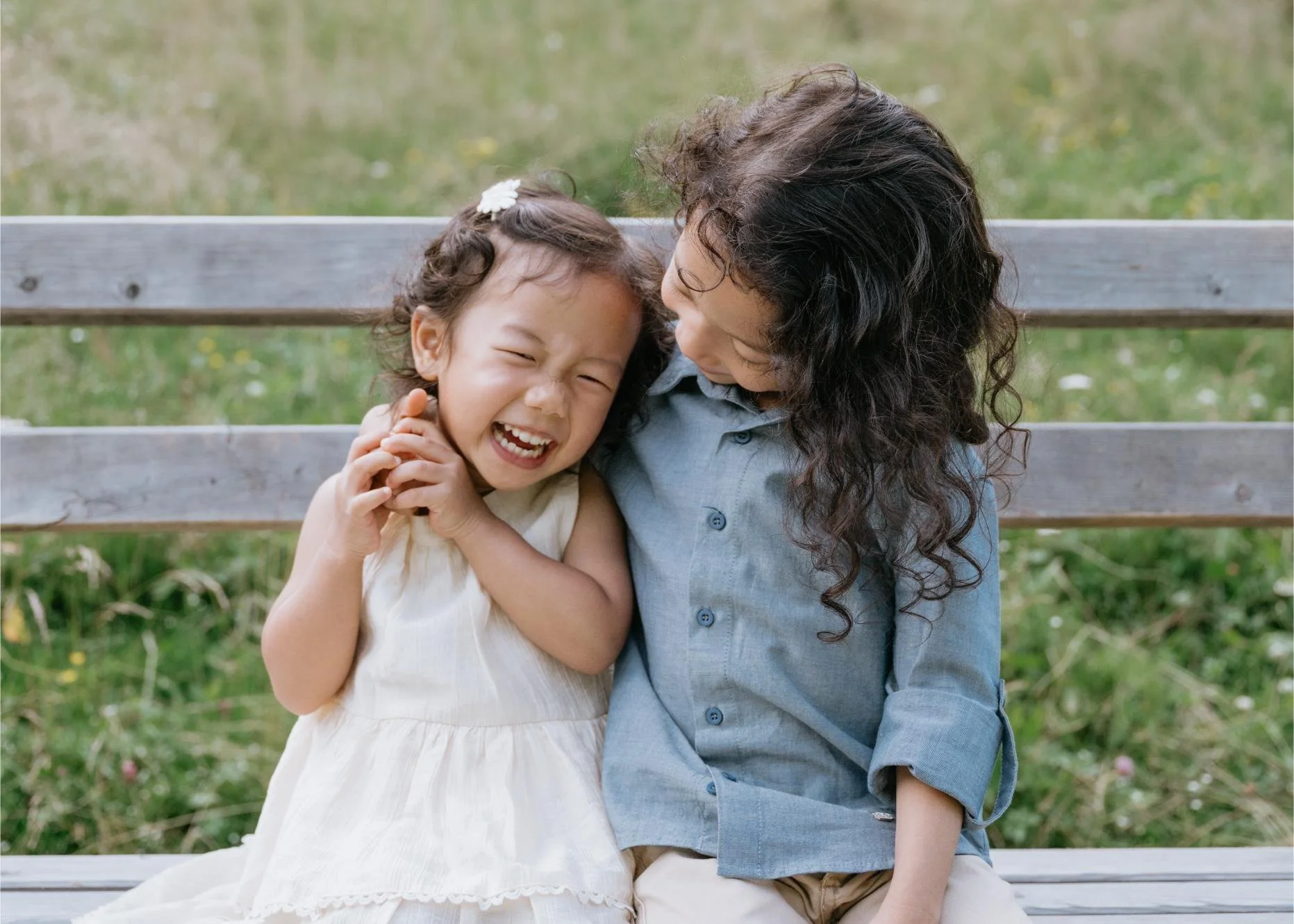 A woman and a young girl sitting on a park bench, playfully holding and laughing at each other.