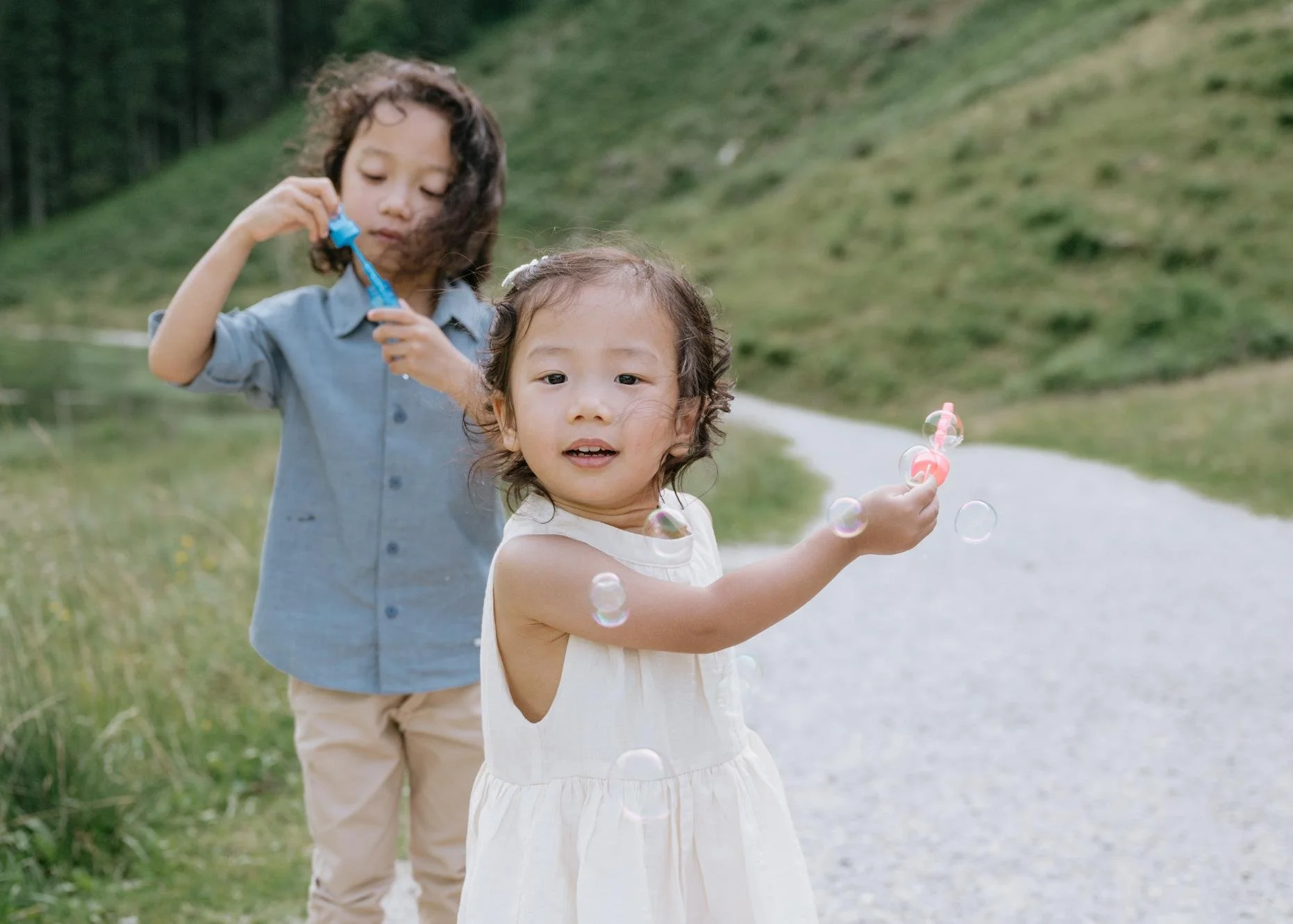 Two children, a girl and a boy, outdoors on a trail, playing with bubbles. The girl is in the foreground, wearing a white dress and holding a bubble wand, while the boy in a blue shirt and beige pants is in the background, also playing with bubbles.