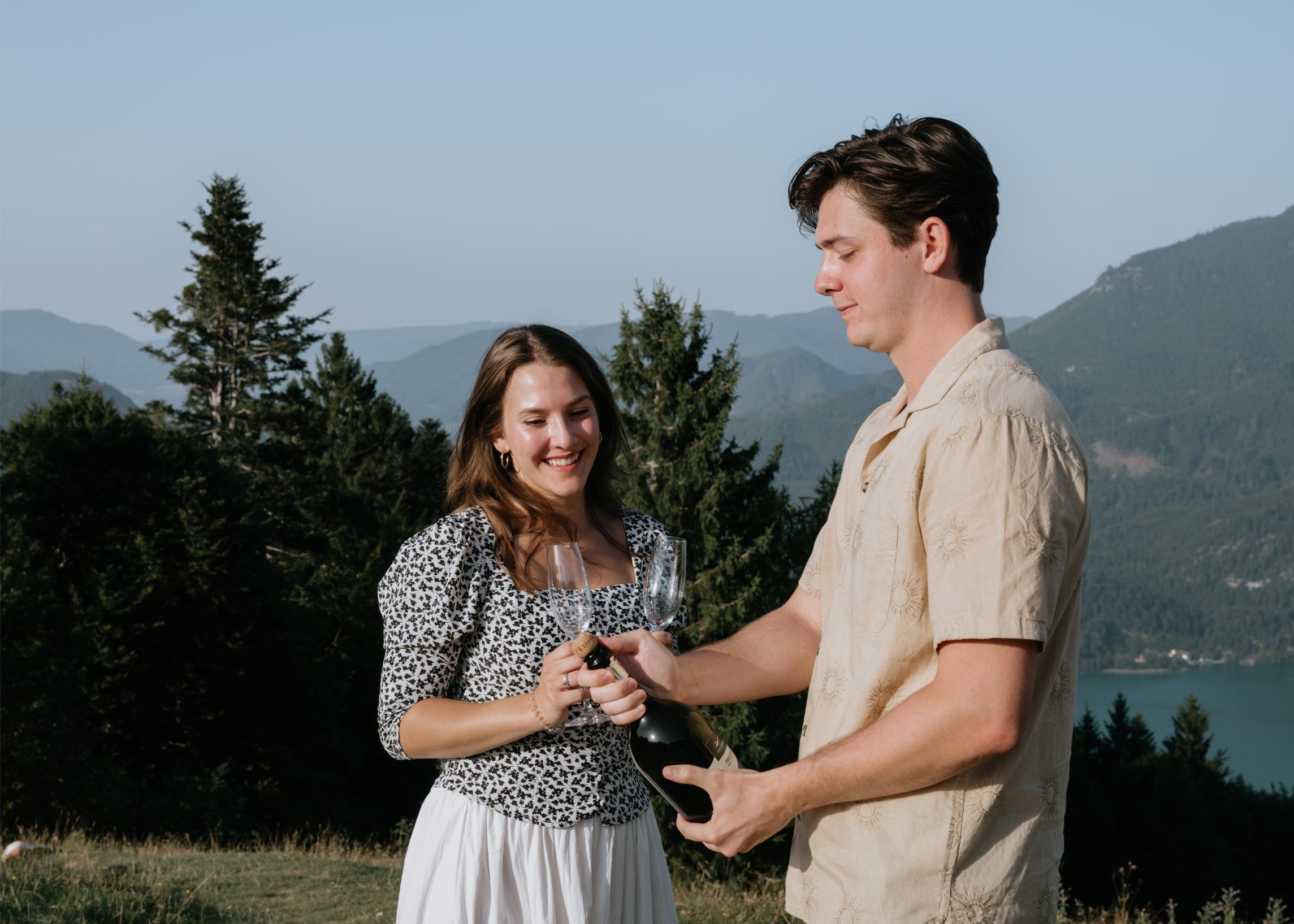 A young couple celebrating outdoors with champagne against a scenic mountain and lake backdrop. The woman is smiling while holding a champagne flute, and the man is opening a bottle of champagne.