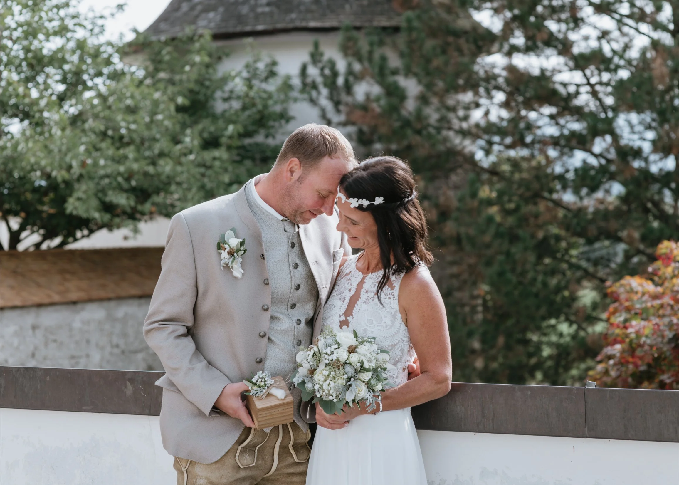 A bride and groom sharing a loving moment outdoors on their wedding day, with trees and a building in the background.