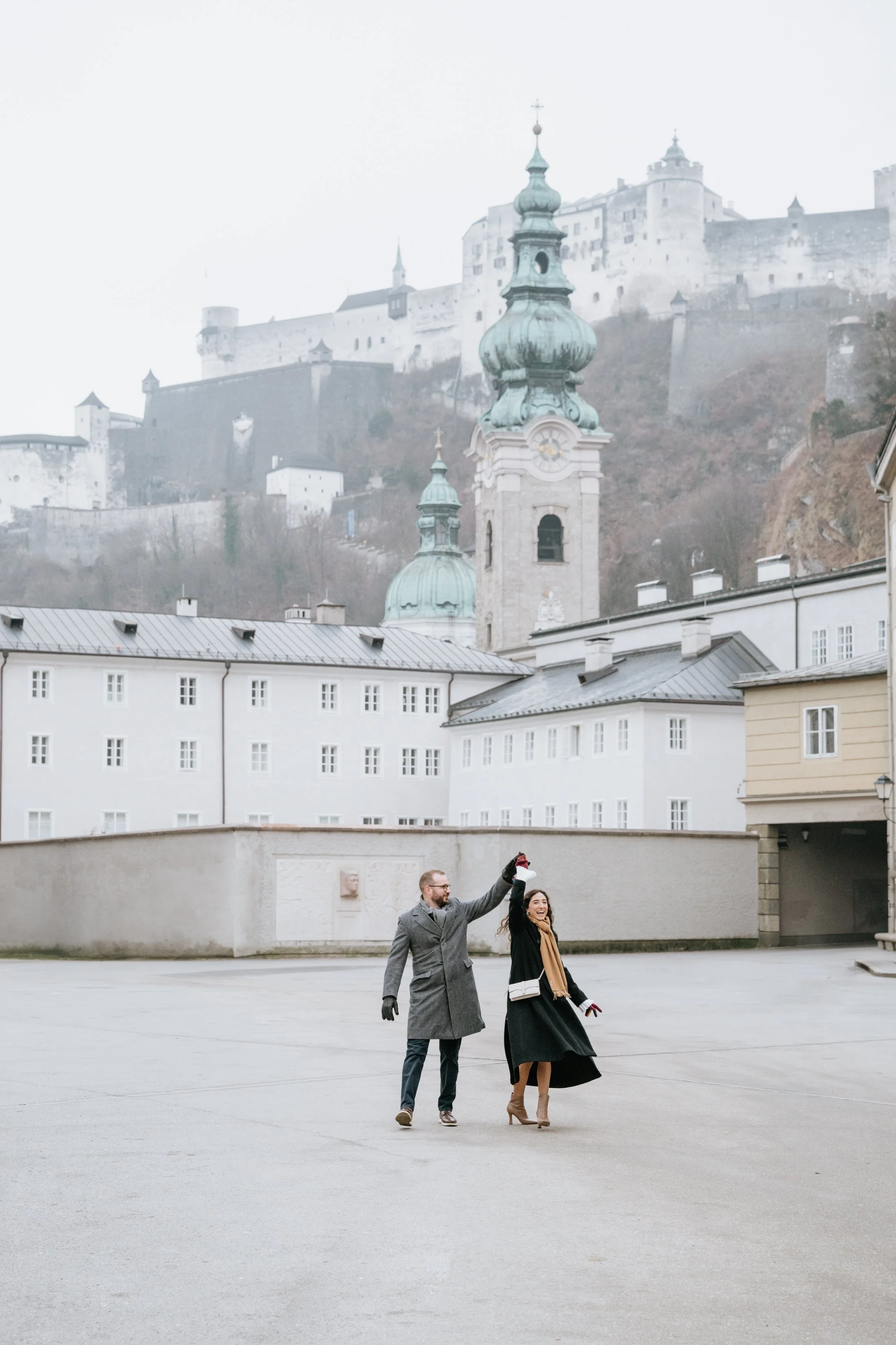 A man and woman holding hands up in front of historic European architecture, including church towers and castle walls, on a cloudy day.