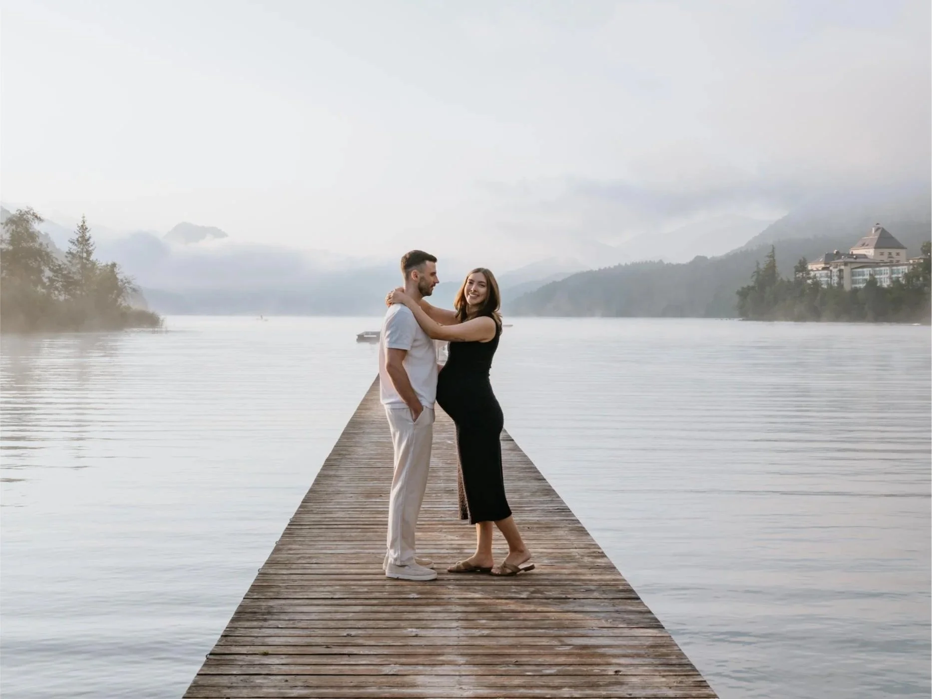 A happy couple standing on a wooden dock by the lake, embracing and smiling, with mountains and buildings in the background.