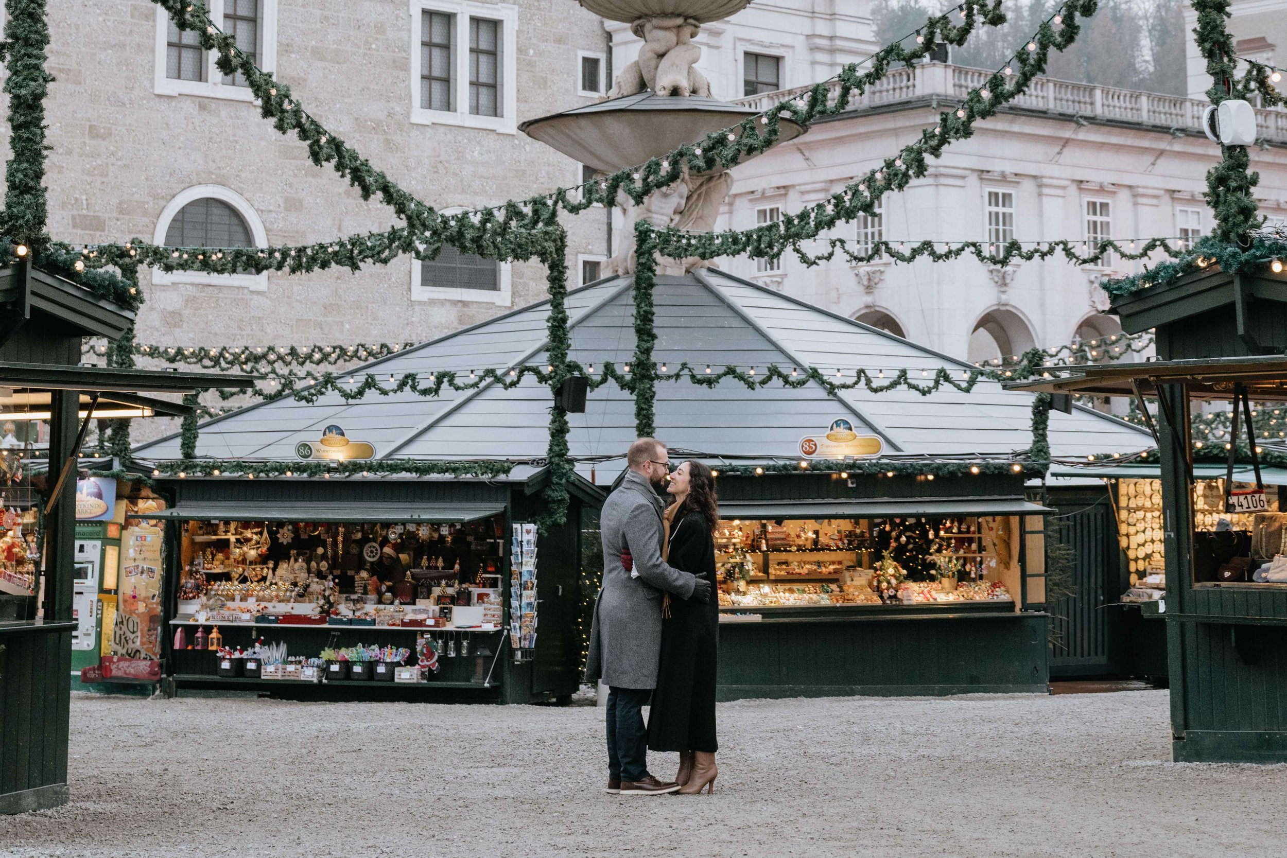 A couple standing closely together, facing each other, at an outdoor holiday market. The market stalls are decorated with Christmas garlands and lights, with a historic building in the background.