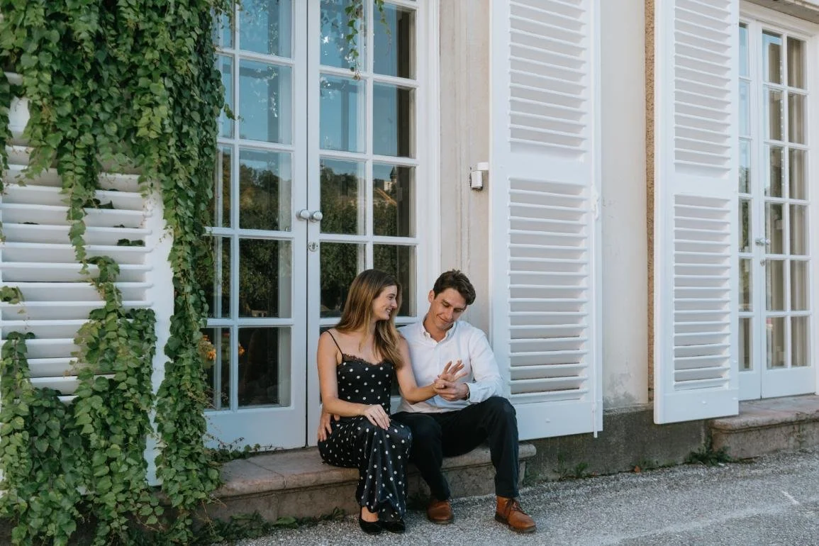 A young couple sitting on a sidewalk outside a house, smiling and looking at a smartphone together. The house has white shutters and large windows, with greenery on the side.
