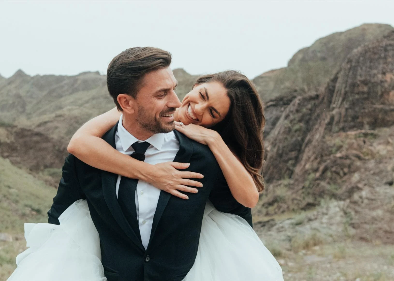 A smiling couple, a man in a tuxedo and a woman in a wedding dress, enjoying a piggyback ride outdoors with mountains in the background.